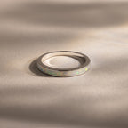 The White Opal Inlay Band in Sterling Silver showcases a shimmering opal inlay set on a sleek silver band, displayed on a beige surface with soft shadows for elegant gemstone appeal.