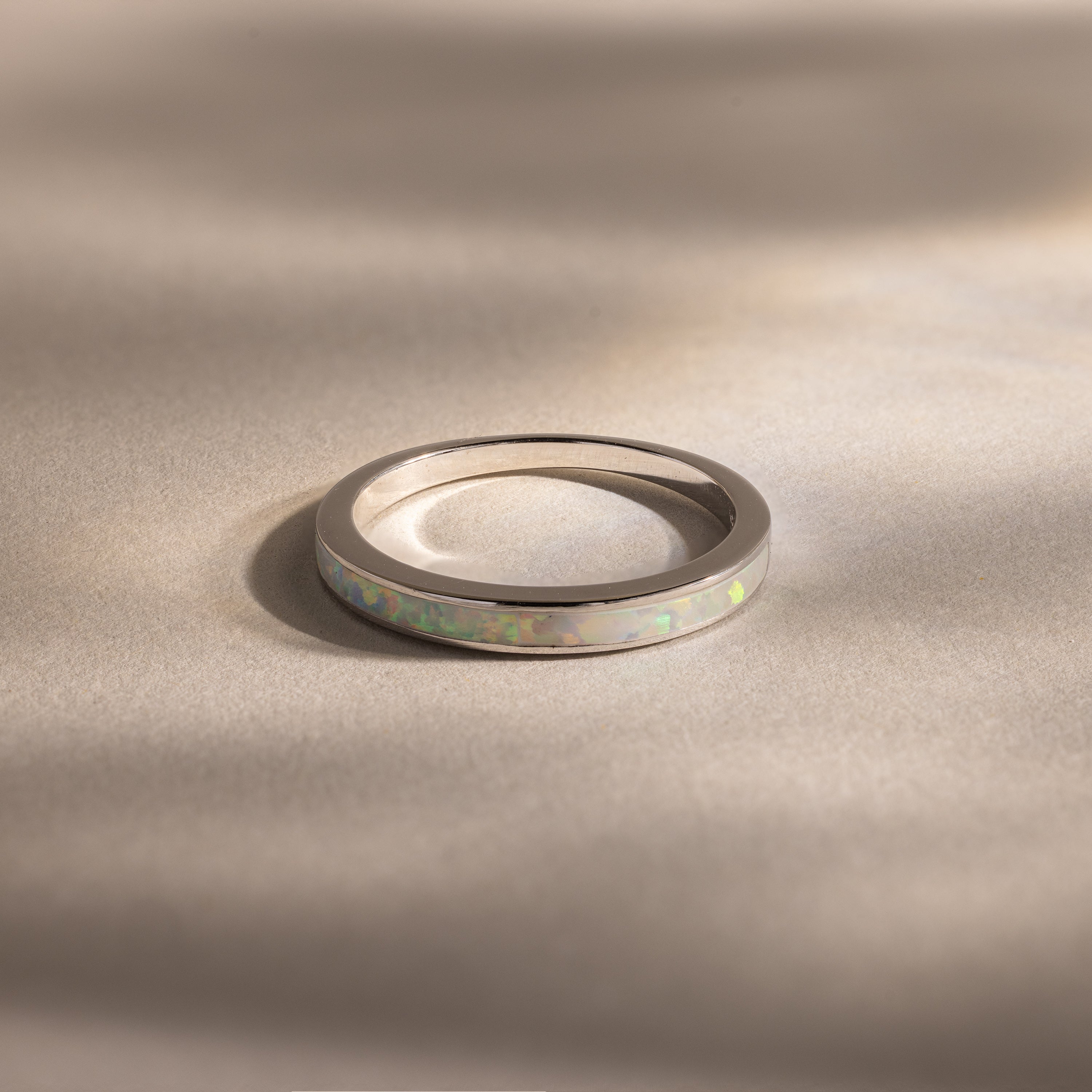 The White Opal Inlay Band in Sterling Silver showcases a shimmering opal inlay set on a sleek silver band, displayed on a beige surface with soft shadows for elegant gemstone appeal.