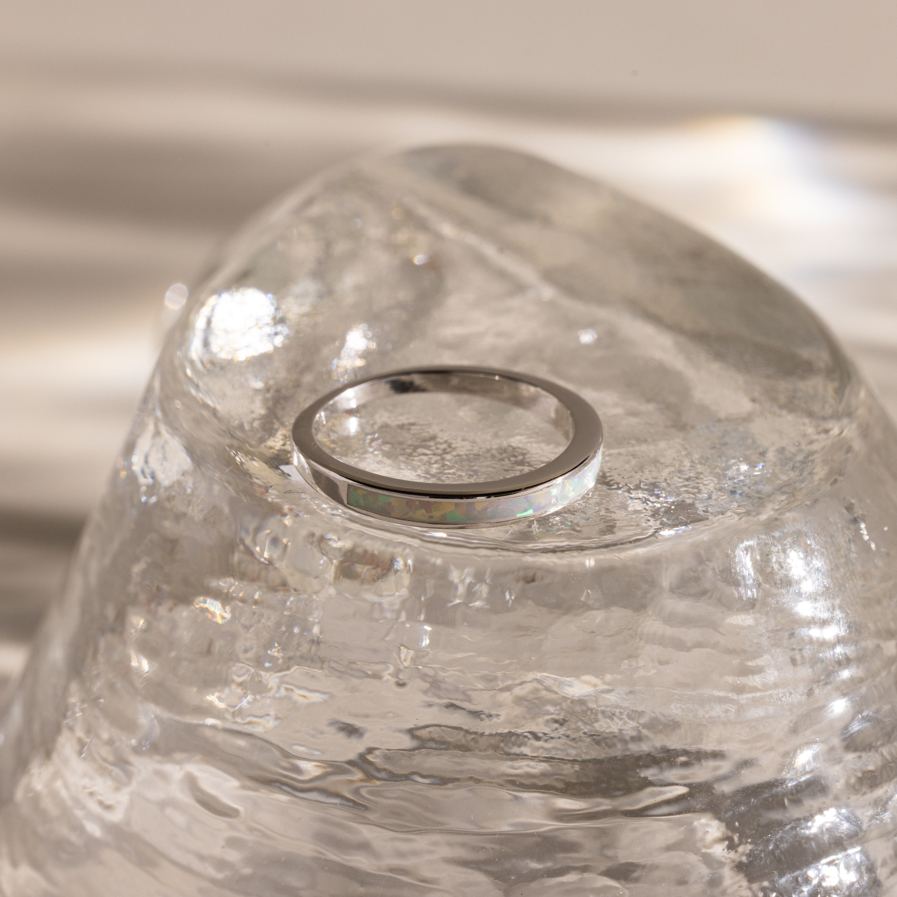 The White Opal Inlay Band in Sterling Silver shines on a clear, textured glass surface, with gentle light reflections enhancing the elegance of this gemstone jewelry.