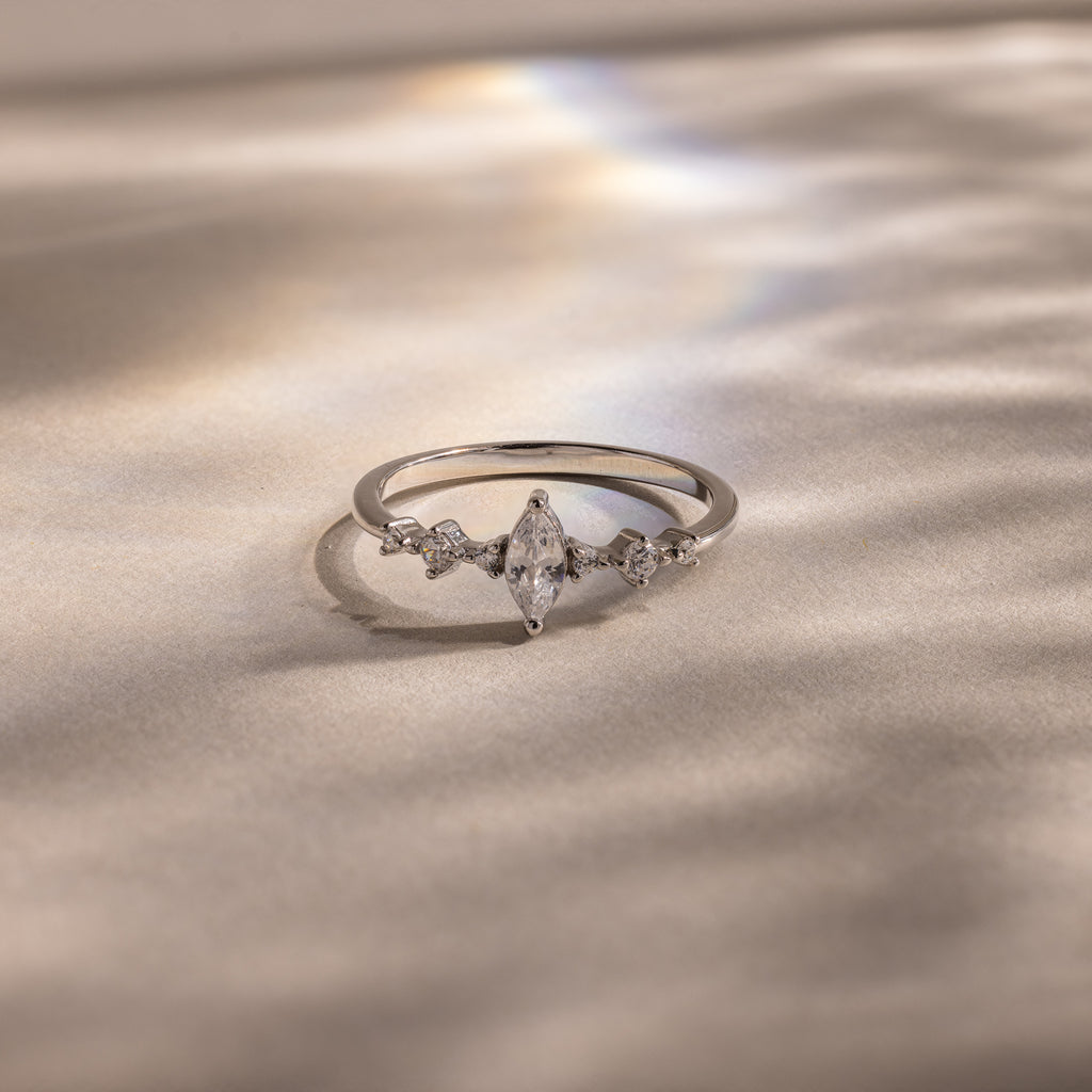 The Harlow Diamond Ring, featuring a marquise diamond with petite side stones set on a silver band, rests on a soft, shimmering surface—an elegant engagement ring choice.