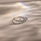 The Harlow Diamond Ring, featuring a marquise diamond with petite side stones set on a silver band, rests on a soft, shimmering surface—an elegant engagement ring choice.