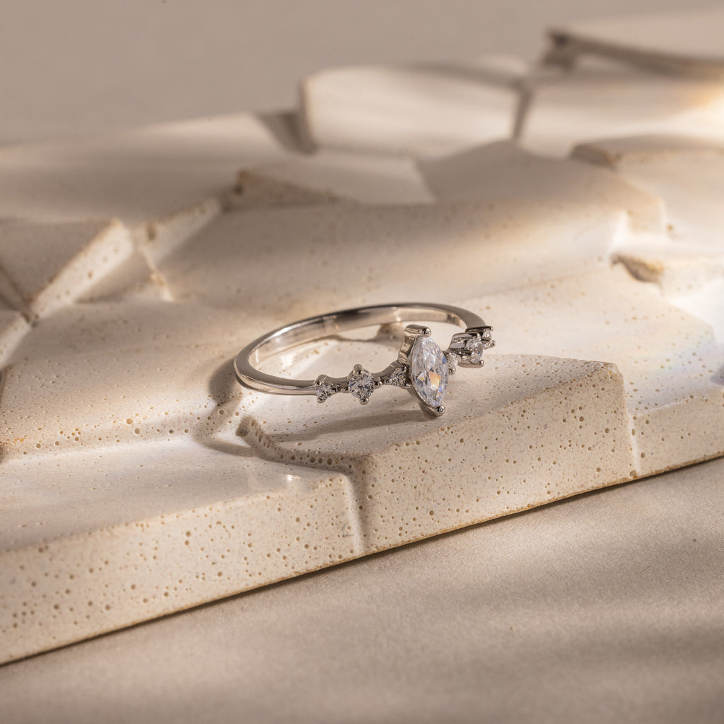 The Harlow Diamond Ring in Sterling Silver, featuring a marquise diamond and delicate side stones, is displayed on a textured beige surface, exuding timeless elegance.