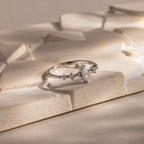 The Harlow Diamond Ring in Sterling Silver, featuring a marquise diamond and delicate side stones, is displayed on a textured beige surface, exuding timeless elegance.