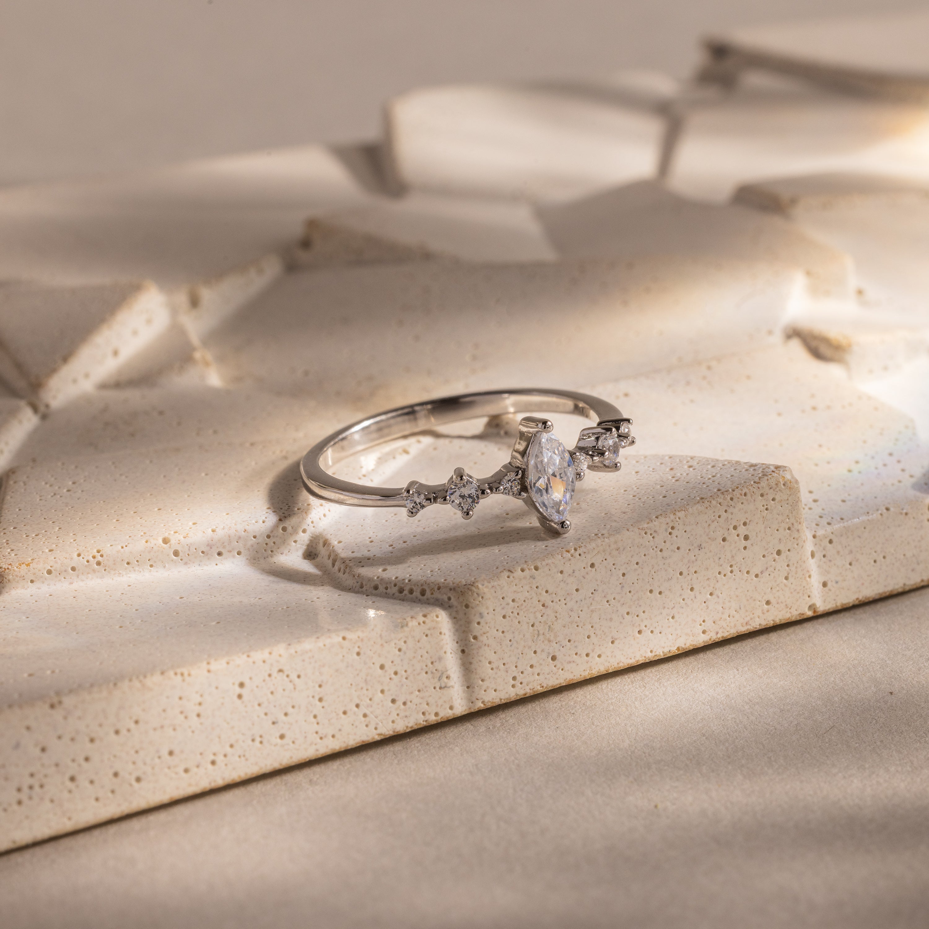 The Harlow Diamond Ring in Sterling Silver, featuring a marquise diamond and delicate side stones, is displayed on a textured beige surface, exuding timeless elegance.