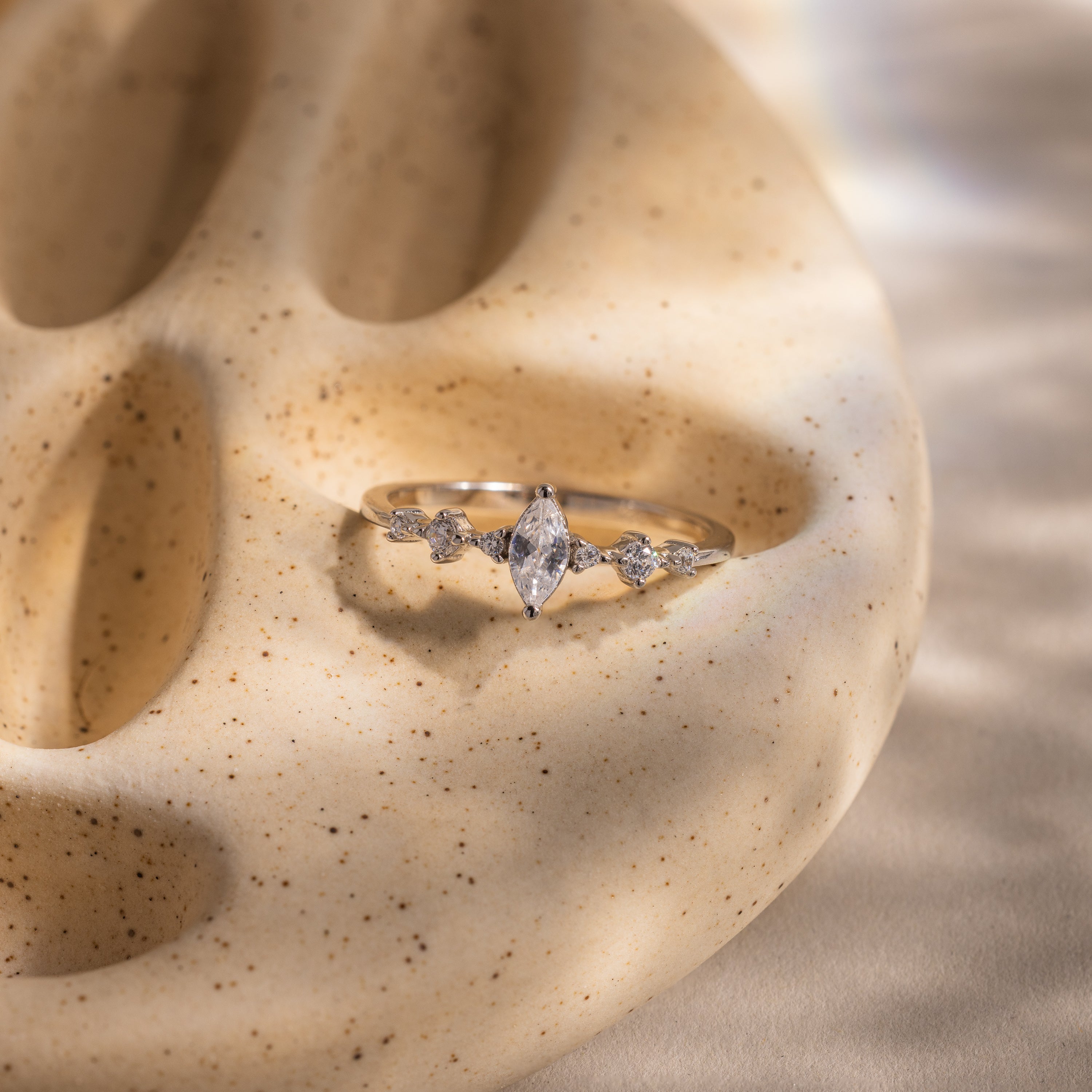 The Harlow Diamond Ring in Sterling Silver, featuring a marquise-cut center and side stones, rests on a textured light beige sculpted surface.