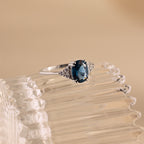 The Elizabeth Blue Topaz Ring in Sterling Silver features an oval blue topaz and small diamonds, styled in a vintage boho design and displayed on a clear textured surface.
