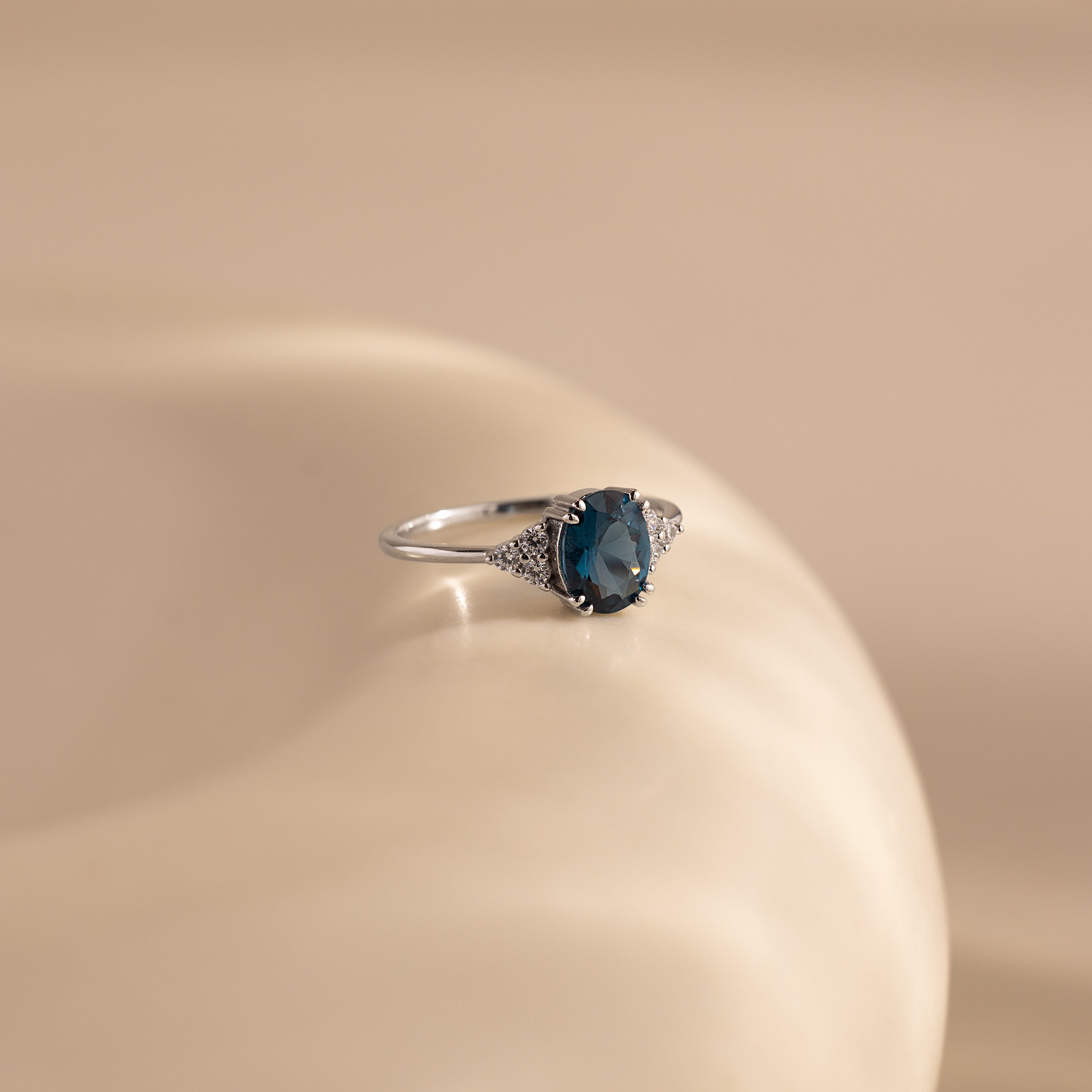 The Elizabeth Blue Topaz Ring in Sterling Silver features an oval blue topaz and small clear stones, displayed on a smooth beige surface—a perfect vintage boho accessory for any occasion.