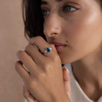 A woman with light brown skin gazes thoughtfully, wearing the Elizabeth Blue Topaz Ring in Sterling Silver—a vintage boho piece featuring a striking blue gemstone on her finger.