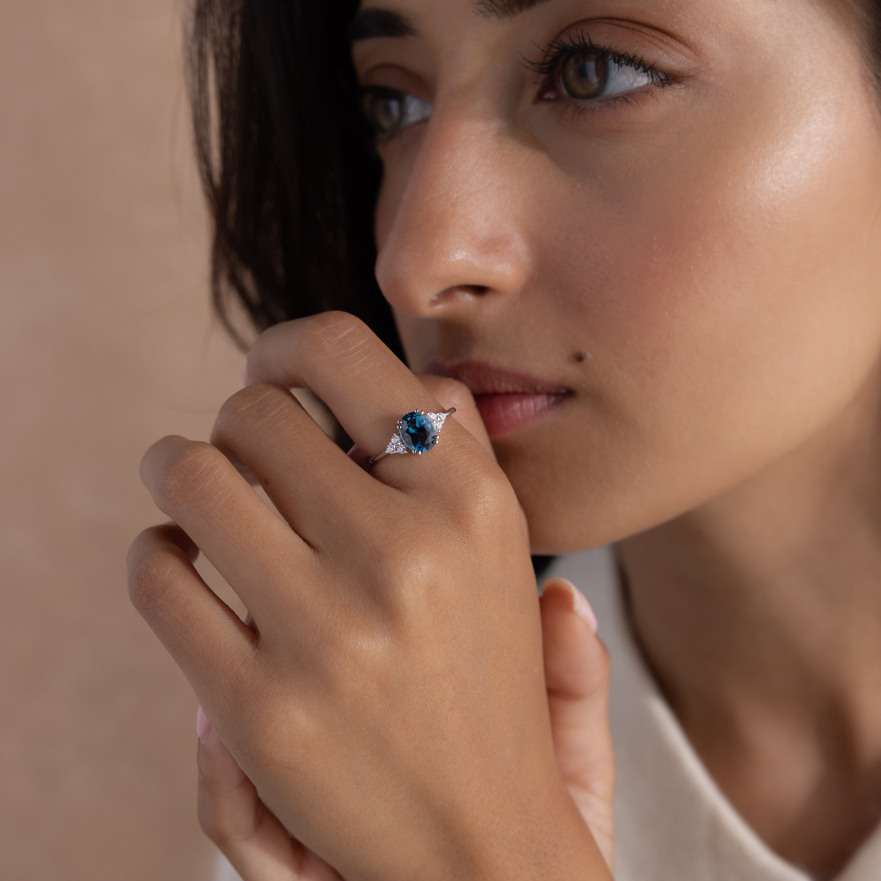 A woman with light brown skin gazes thoughtfully, wearing the Elizabeth Blue Topaz Ring in Sterling Silver—a vintage boho piece featuring a striking blue gemstone on her finger.