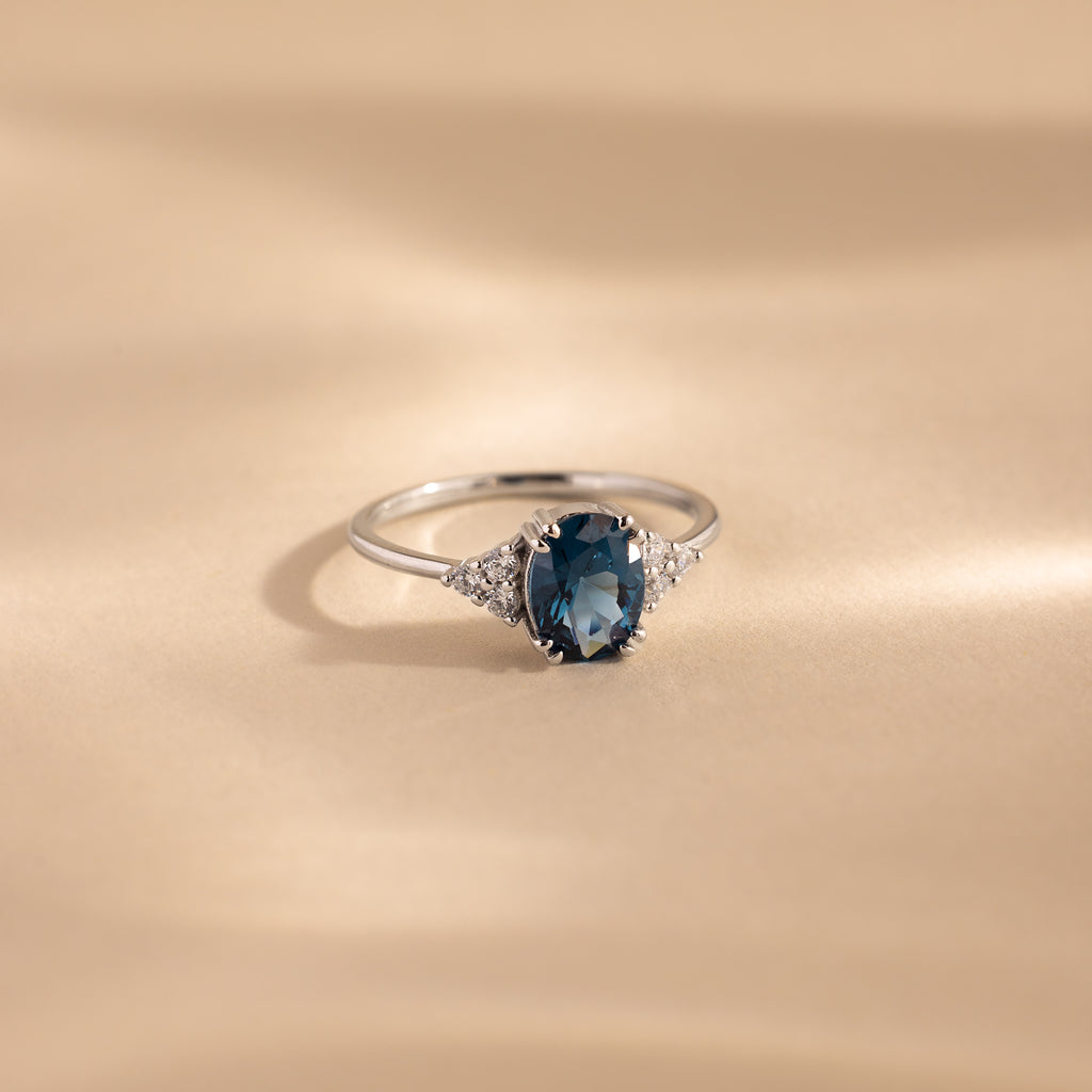 A silver ring with an oval-cut deep blue topaz center stone, flanked by clusters of sparkling white accent stones, displayed elegantly on a softly lit beige surface.