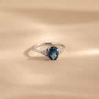 A silver ring with an oval-cut deep blue topaz center stone, flanked by clusters of sparkling white accent stones, displayed elegantly on a softly lit beige surface.