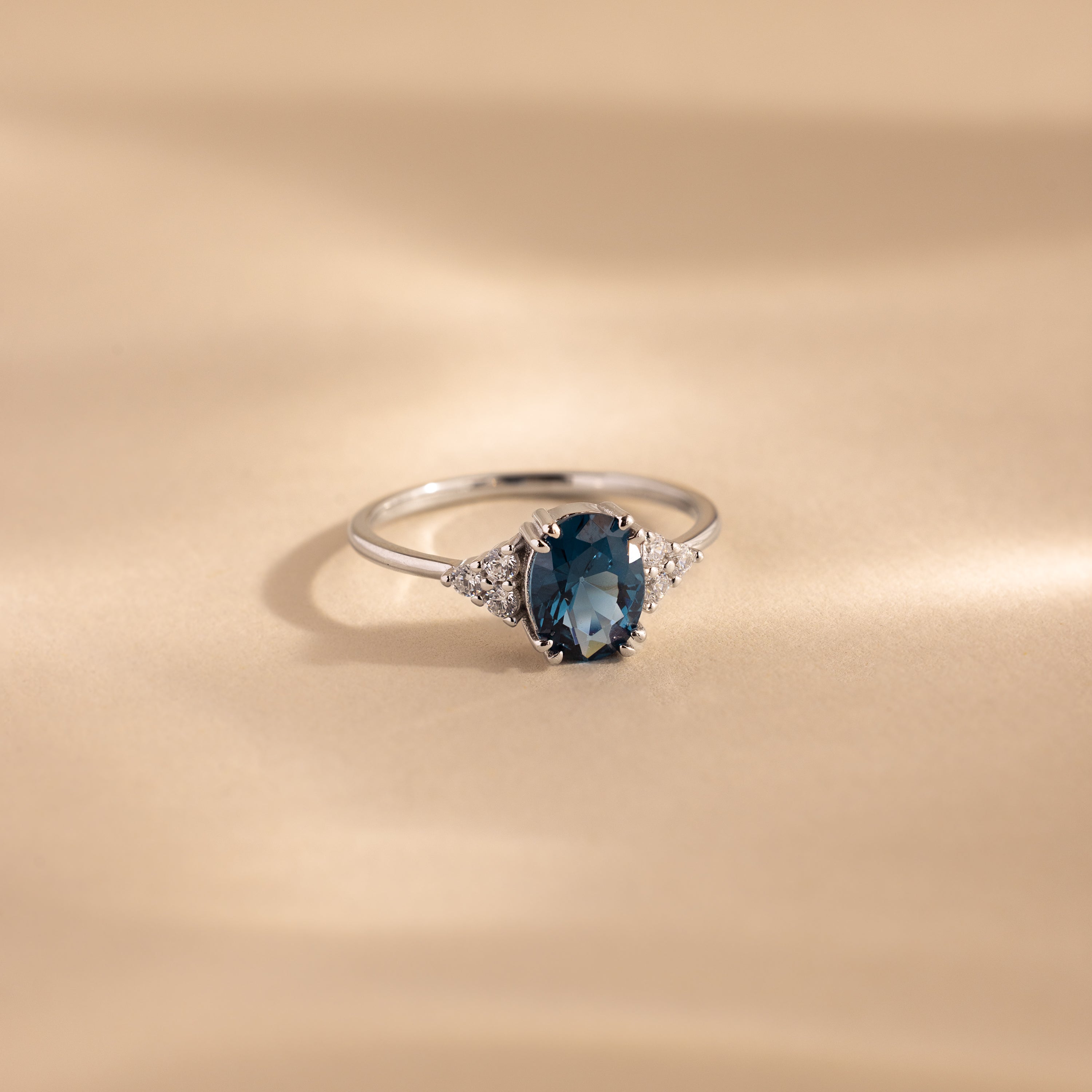A silver ring with an oval-cut deep blue topaz center stone, flanked by clusters of sparkling white accent stones, displayed elegantly on a softly lit beige surface.