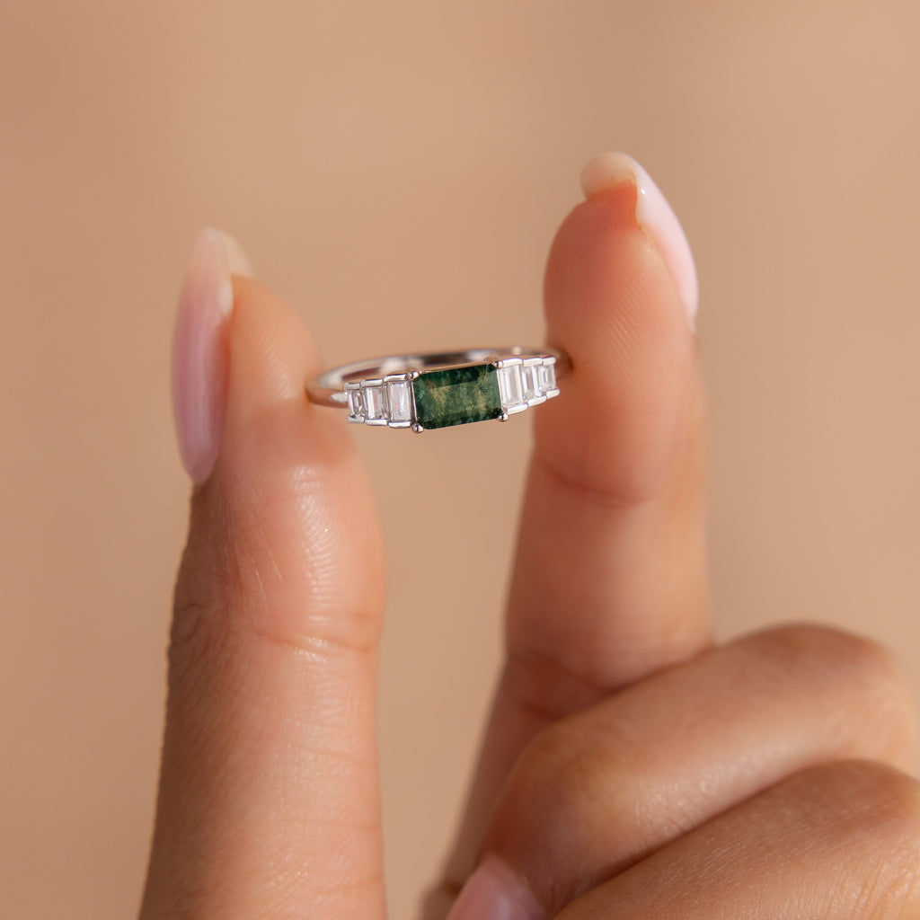 The ring is held delicately between two fingers against a soft peach-toned background. The elevated view showcases the symmetrical stone setting and the glossy finish of the sterling silver.