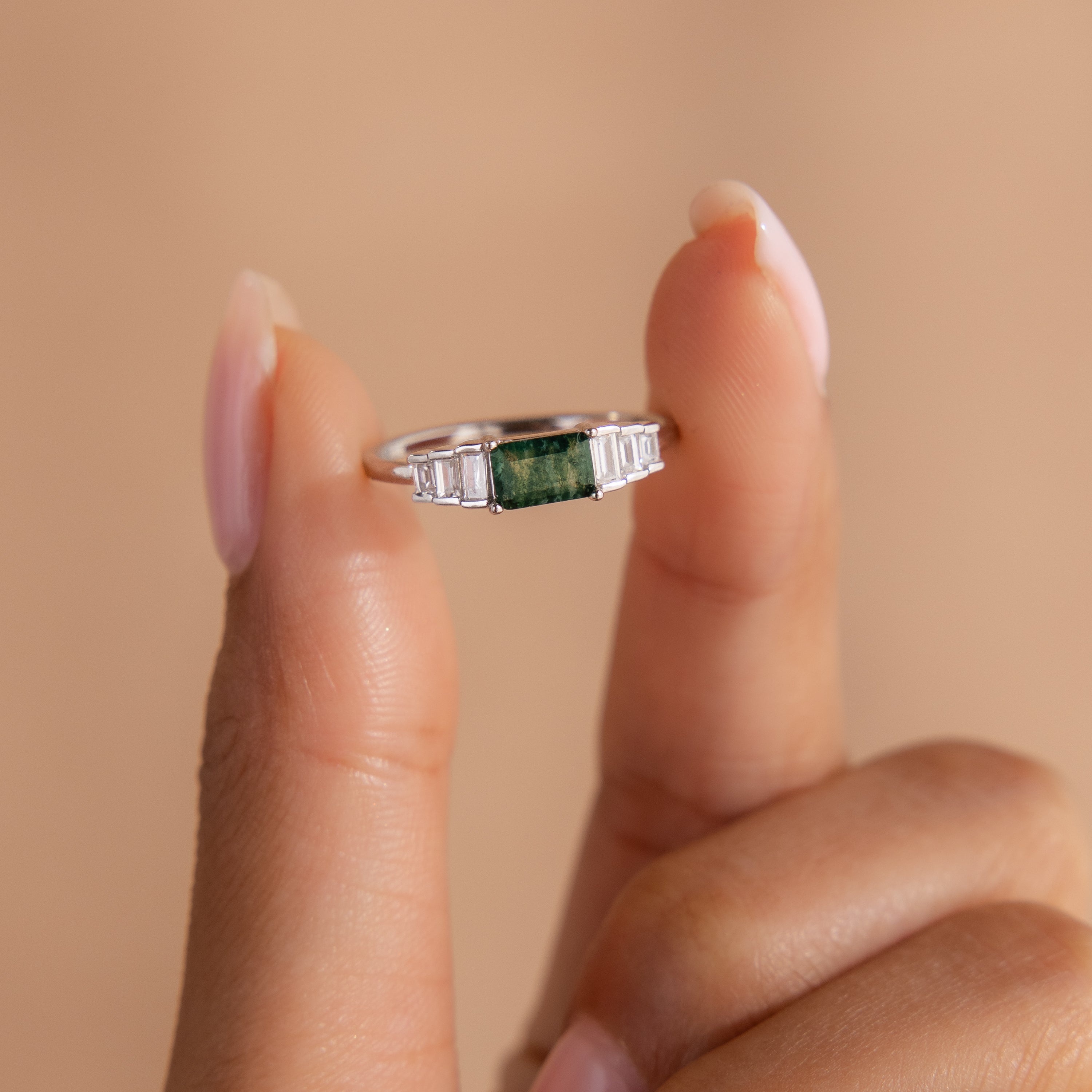 The ring is held delicately between two fingers against a soft peach-toned background. The elevated view showcases the symmetrical stone setting and the glossy finish of the sterling silver.