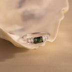 A close-up shot of a sterling silver ring resting atop a seashell, showcasing a rectangular moss agate center stone with intricate green inclusions. The gemstone is flanked by a row of vertically set baguette-cut clear stones on each side, all held in a polished silver band. Soft natural lighting highlights the textures of the shell and the subtle sparkle of the stones.
