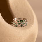 The Emerald Stacking Ring Set in Sterling Silver features vintage-inspired design with green and clear gemstones, elegantly displayed on a smooth beige surface.