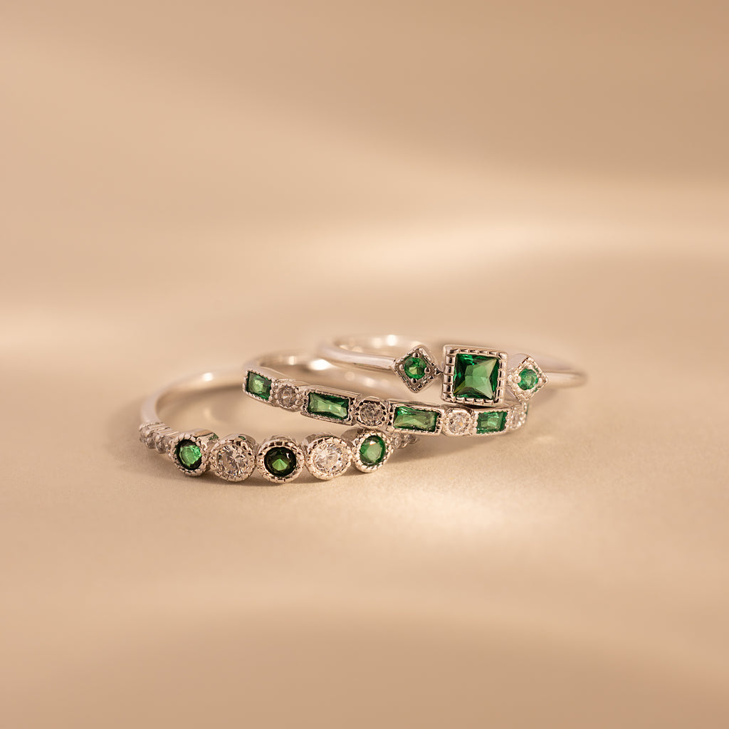 The Emerald Stacking Ring Set features three silver rings with green gemstones and diamond accents, offering vintage-inspired charm for an elegant layered look on a soft beige background.