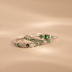 The Emerald Stacking Ring Set features three silver rings with green gemstones and diamond accents, offering vintage-inspired charm for an elegant layered look on a soft beige background.