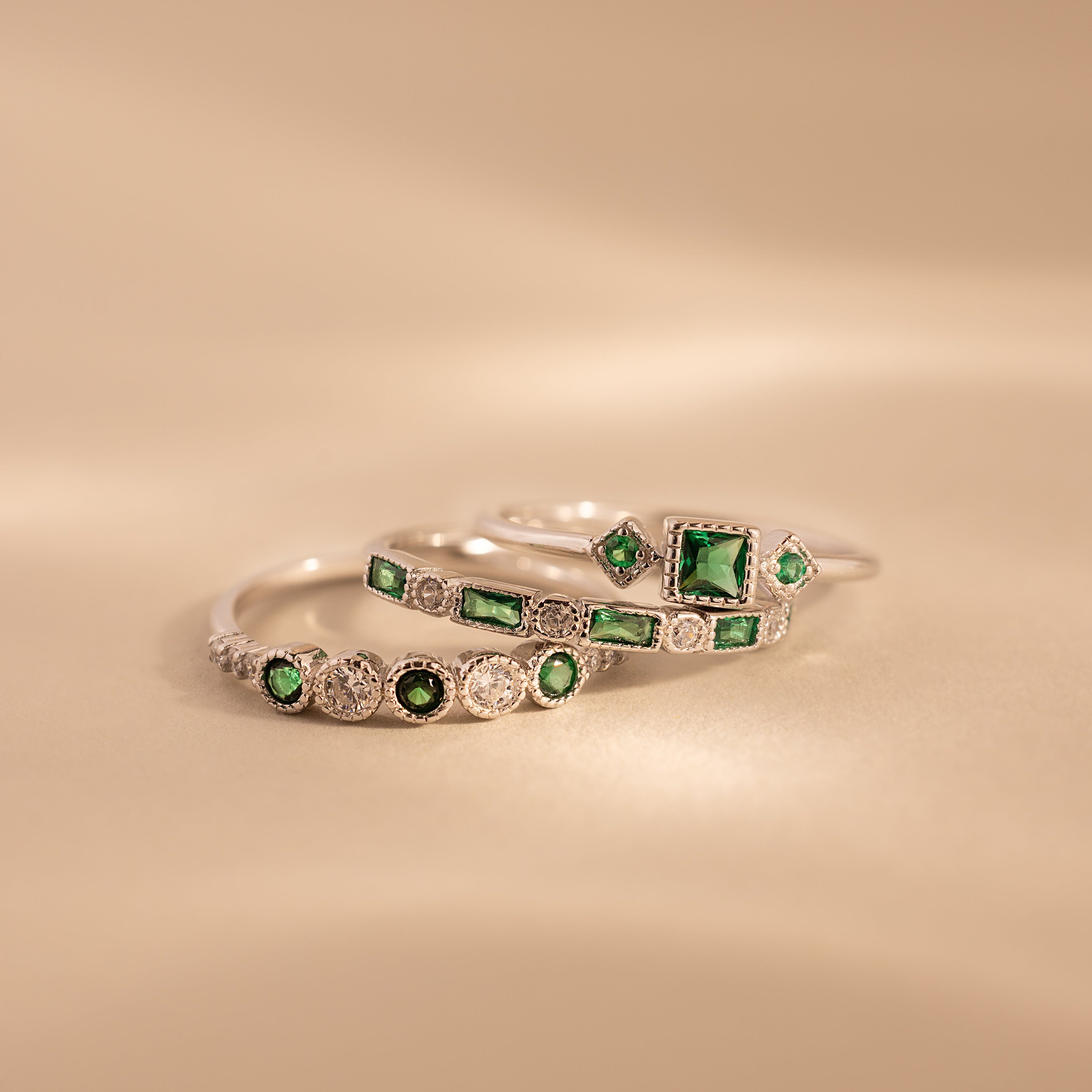 The Emerald Stacking Ring Set features three silver rings with green gemstones and diamond accents, offering vintage-inspired charm for an elegant layered look on a soft beige background.