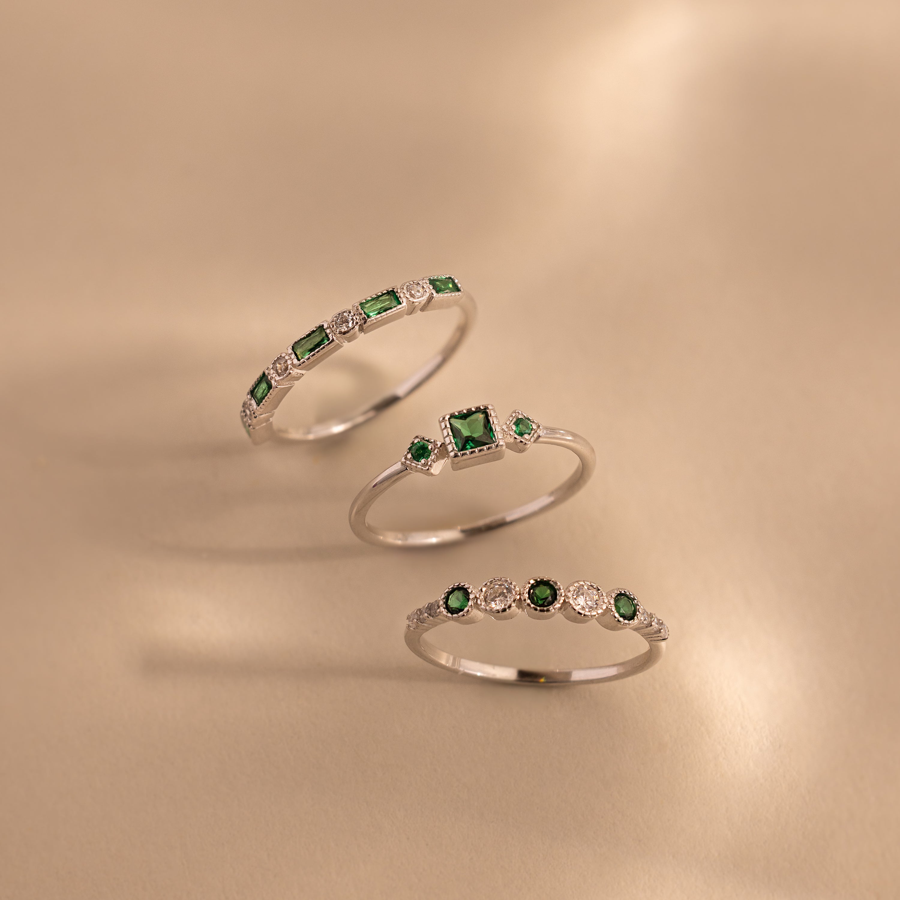 Three sterling silver rings from the Emerald Stacking Ring Set are displayed on a beige surface, showcasing green and white gemstones and offering a vintage-inspired art deco jewelry look.