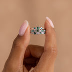 A hand with light pink nails displays the Emerald Stacking Ring Set in Sterling Silver, featuring green and clear gemstones in a vintage-inspired art deco style against a beige background.