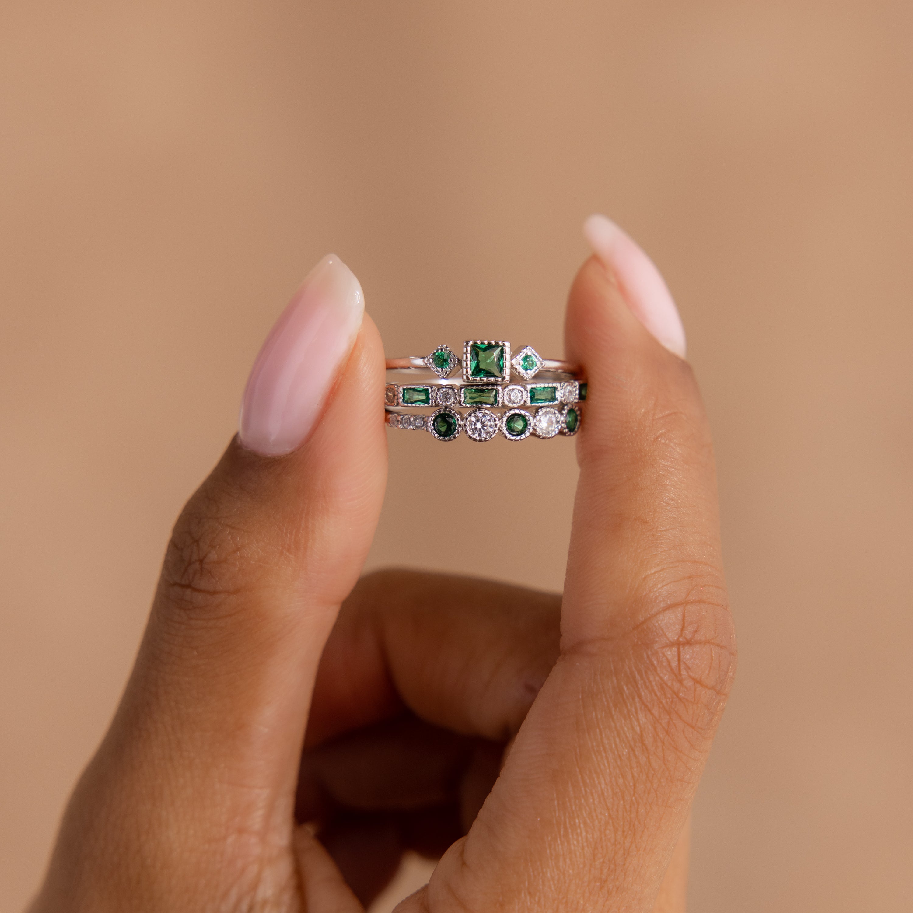 A hand with light pink nails displays the Emerald Stacking Ring Set in Sterling Silver, featuring green and clear gemstones in a vintage-inspired art deco style against a beige background.