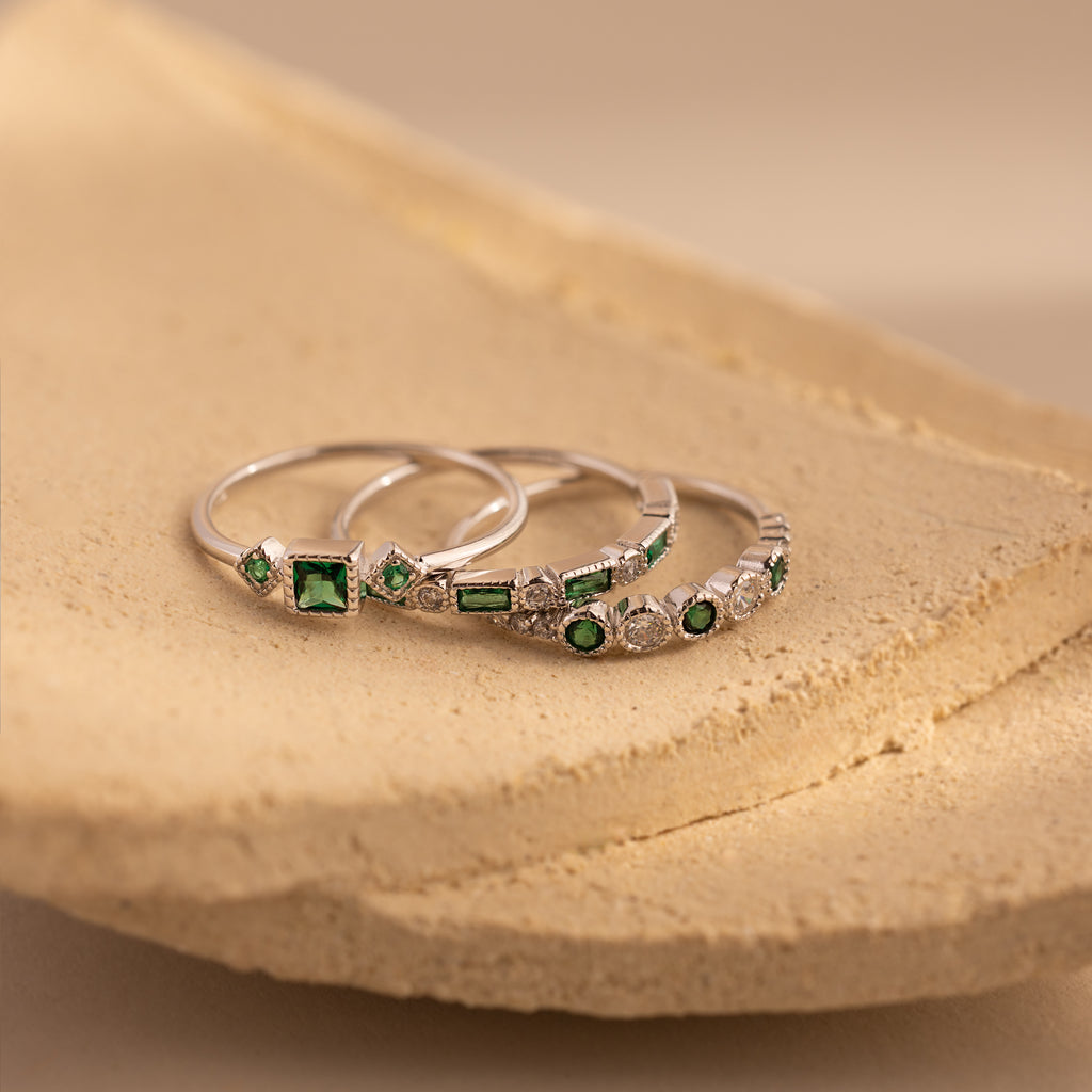 The Emerald Stacking Ring Set in Sterling Silver, featuring green gemstones and diamonds, is displayed on a textured beige surface—a perfect touch of vintage-inspired Art Deco jewelry.