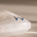 Blue Zircon Flatback Studs featuring blue gemstone flowers are displayed on a smooth, white stone surface.