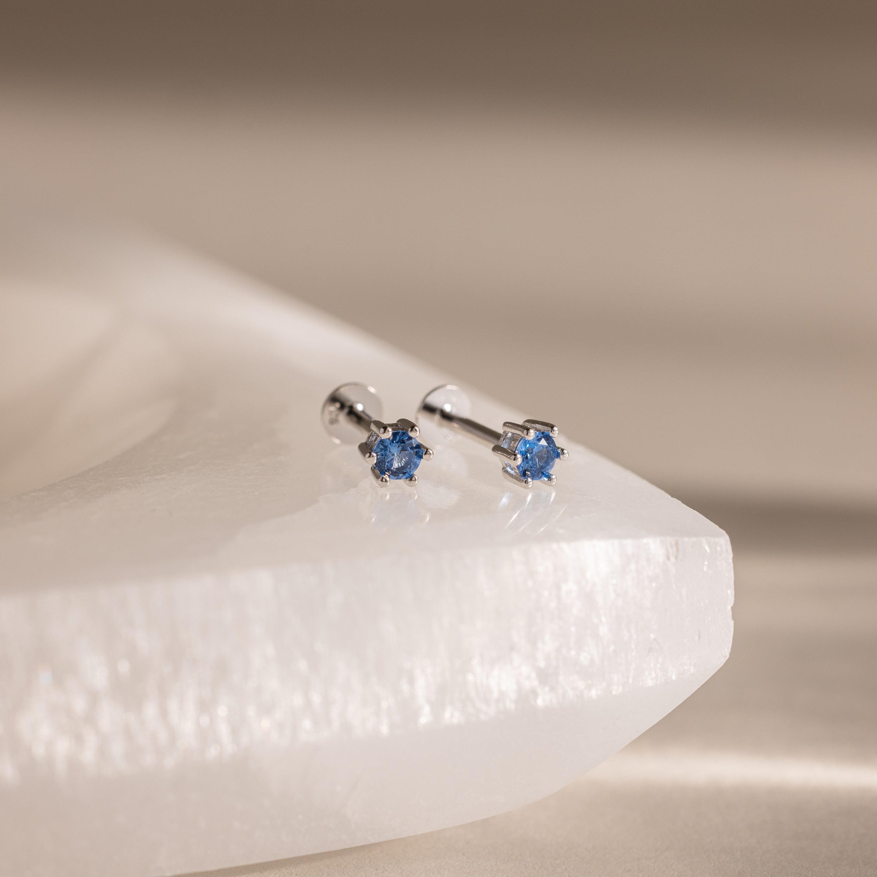 Blue Zircon Flatback Studs featuring blue gemstone flowers are displayed on a smooth, white stone surface.
