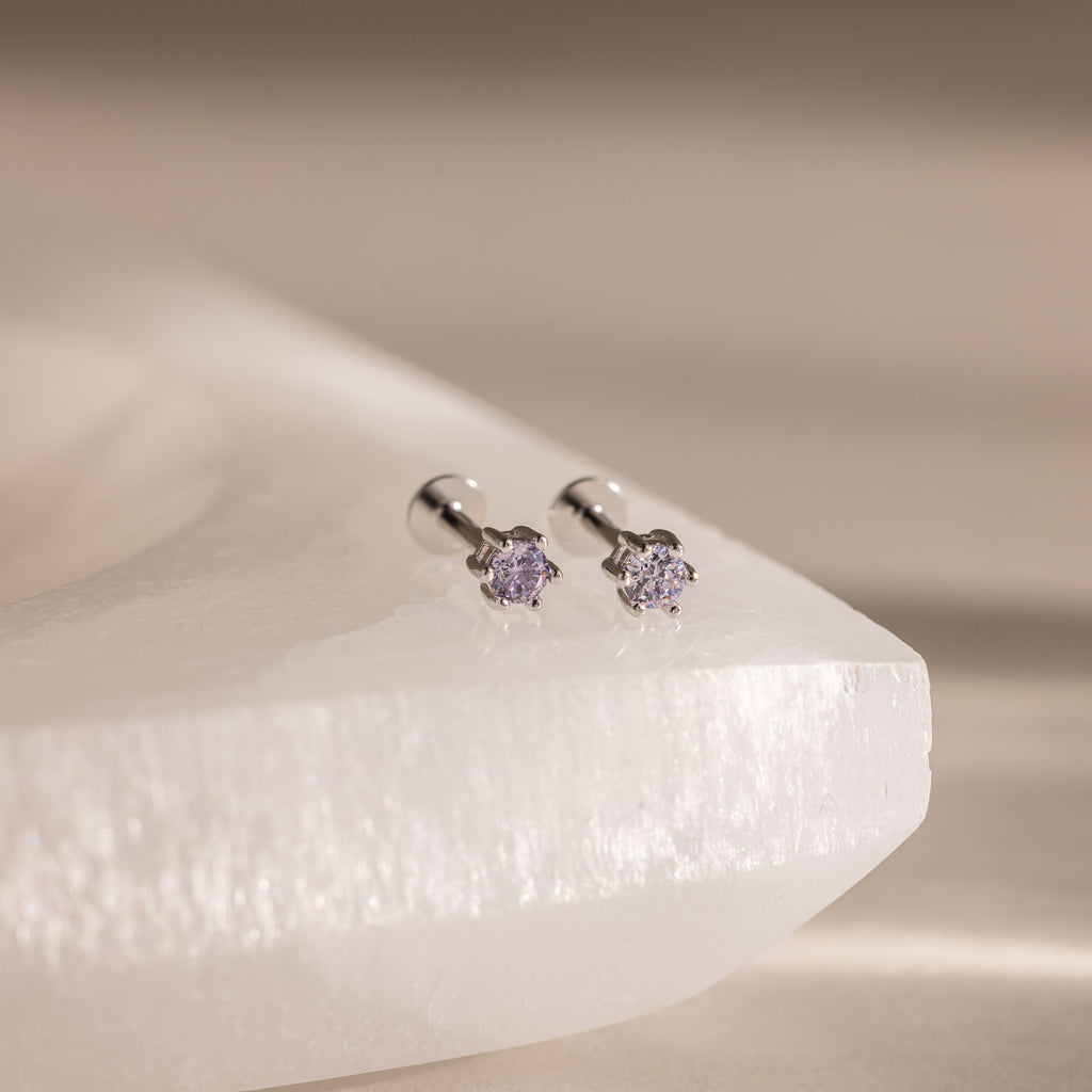 A pair of Alexandrite Flatback Studs with small purple gemstones is displayed on a white stone surface.