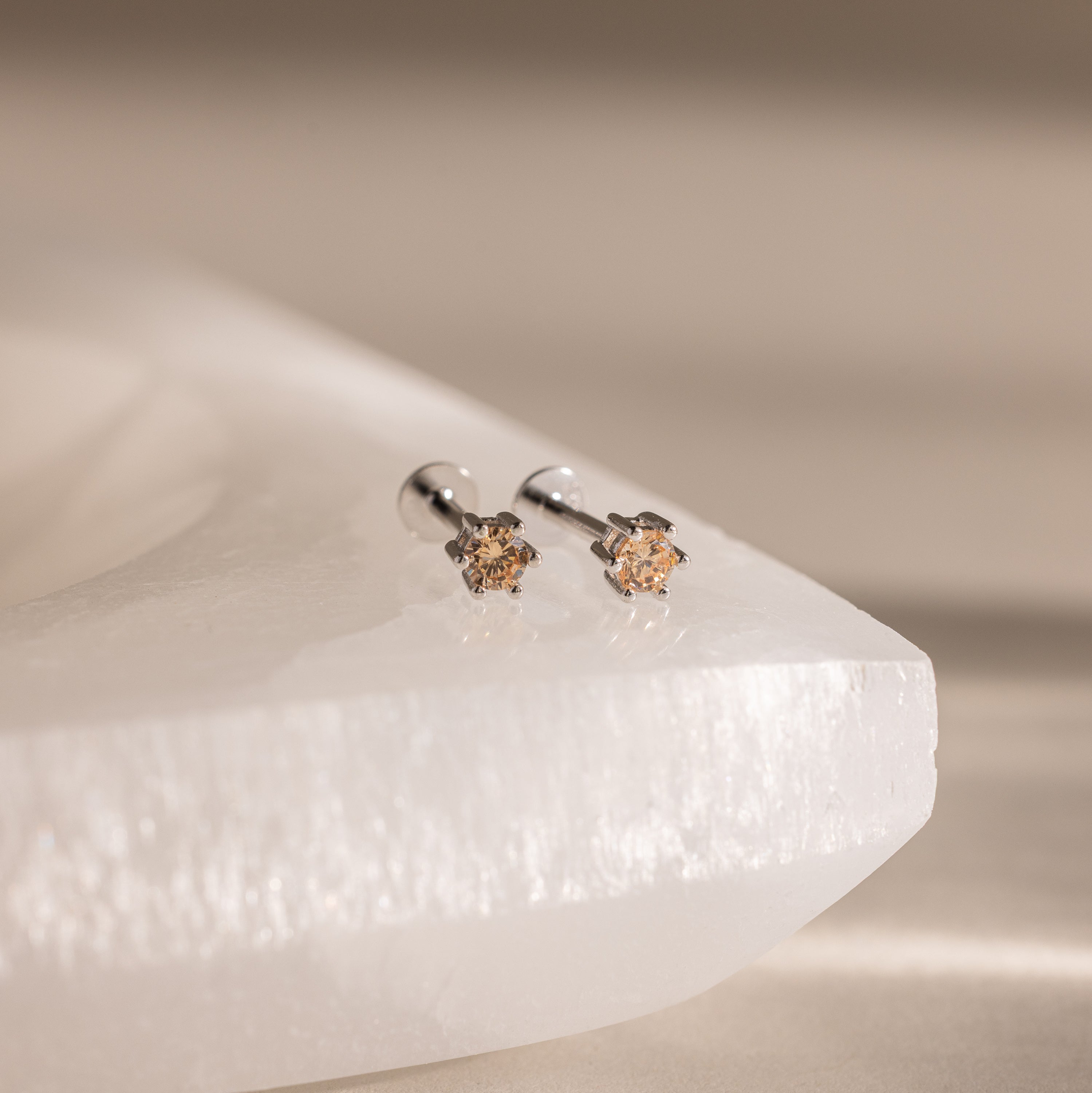 Topaz Flatback Studs, featuring small, round, light brown gemstones on silver, rest on a white textured surface—ideal as birthstone studs for anyone seeking personalized jewelry with subtle elegance.
