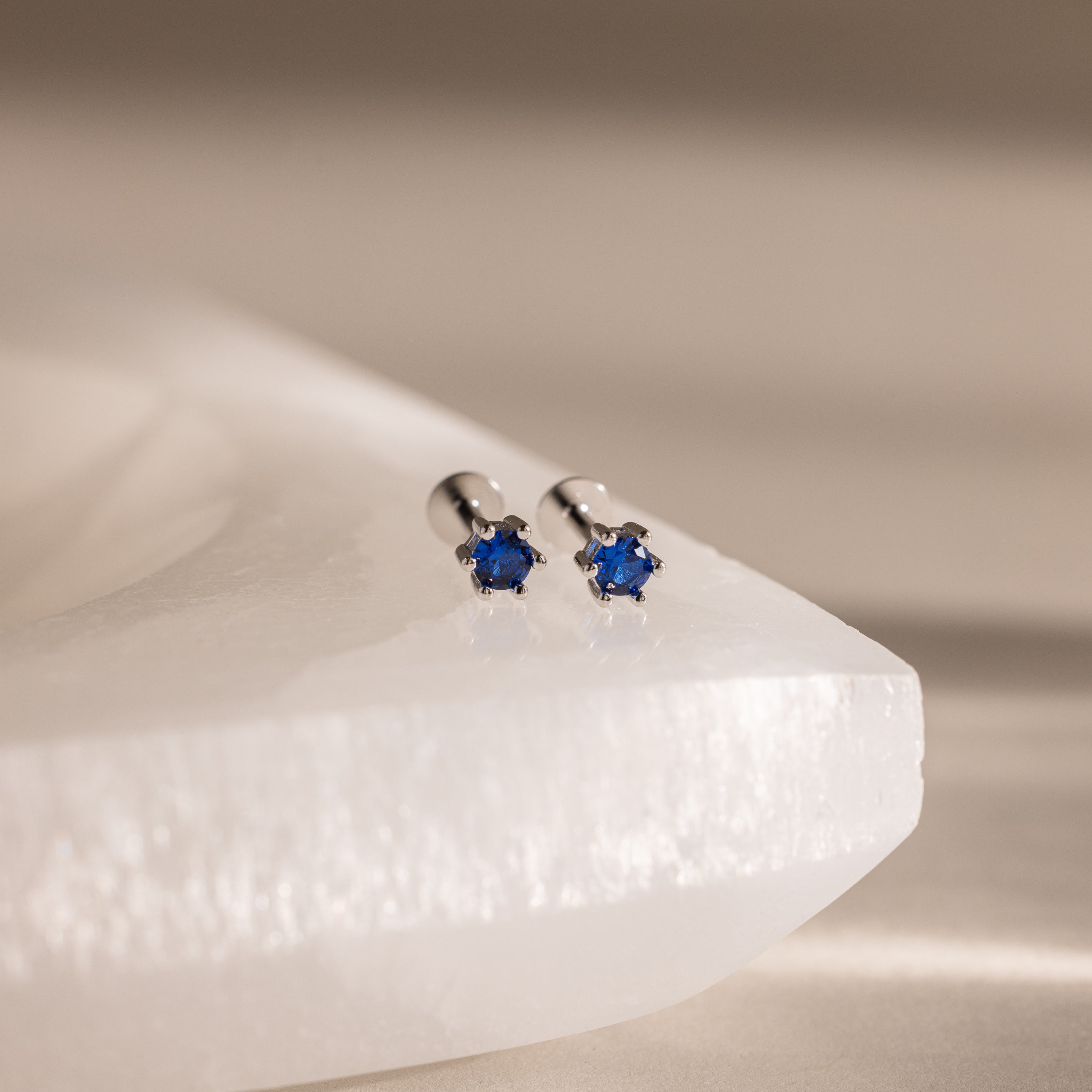 Sapphire Flatback Studs—silver studs with small round blue gemstones, shown on a white textured surface. Elegant birthstone jewelry, perfect for celebrating your birth month.