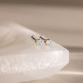 Two Opal Flatback Studs with silver settings rest on a smooth, white stone surface—an ideal choice for personalized jewelry lovers.