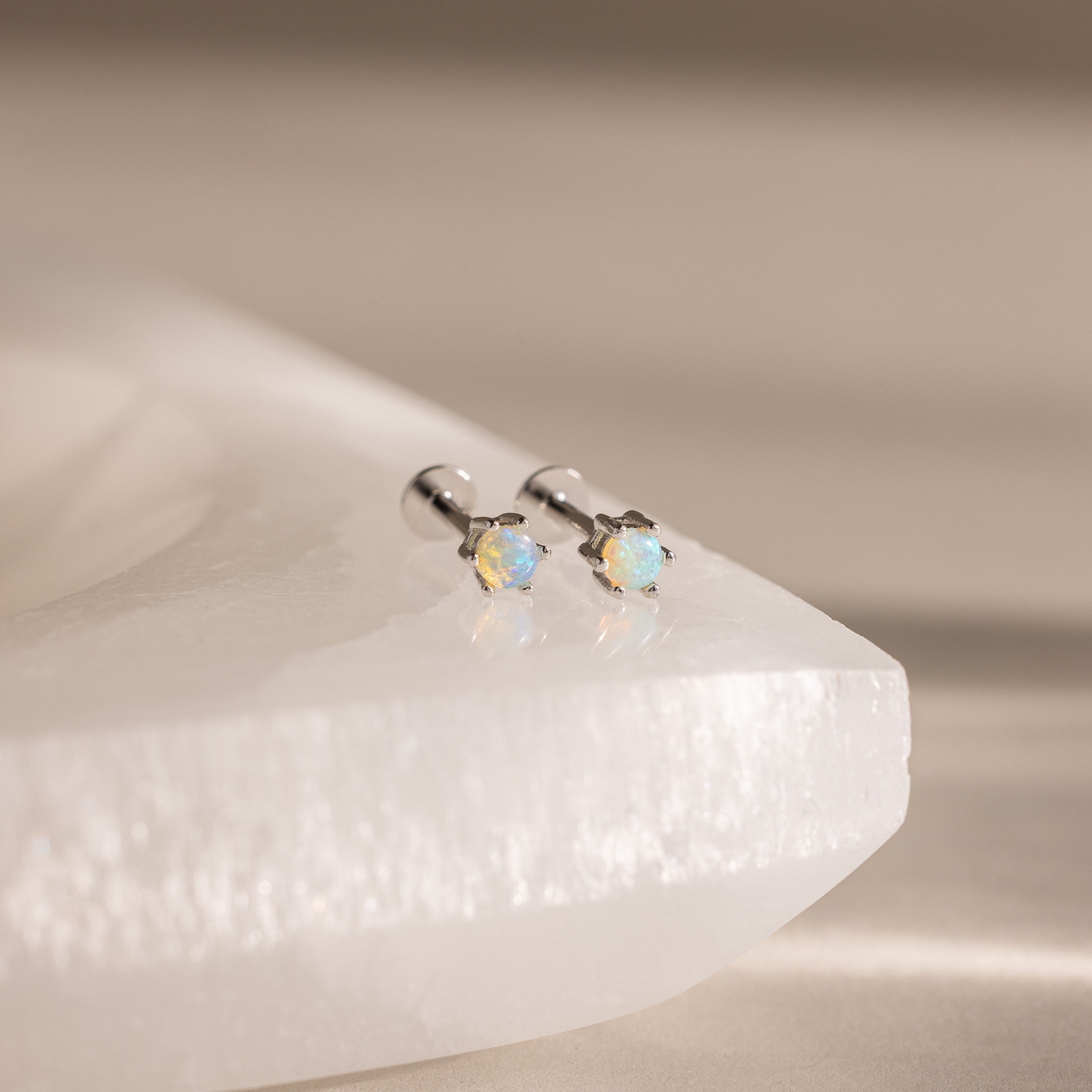 Two Opal Flatback Studs with silver settings rest on a smooth, white stone surface—an ideal choice for personalized jewelry lovers.