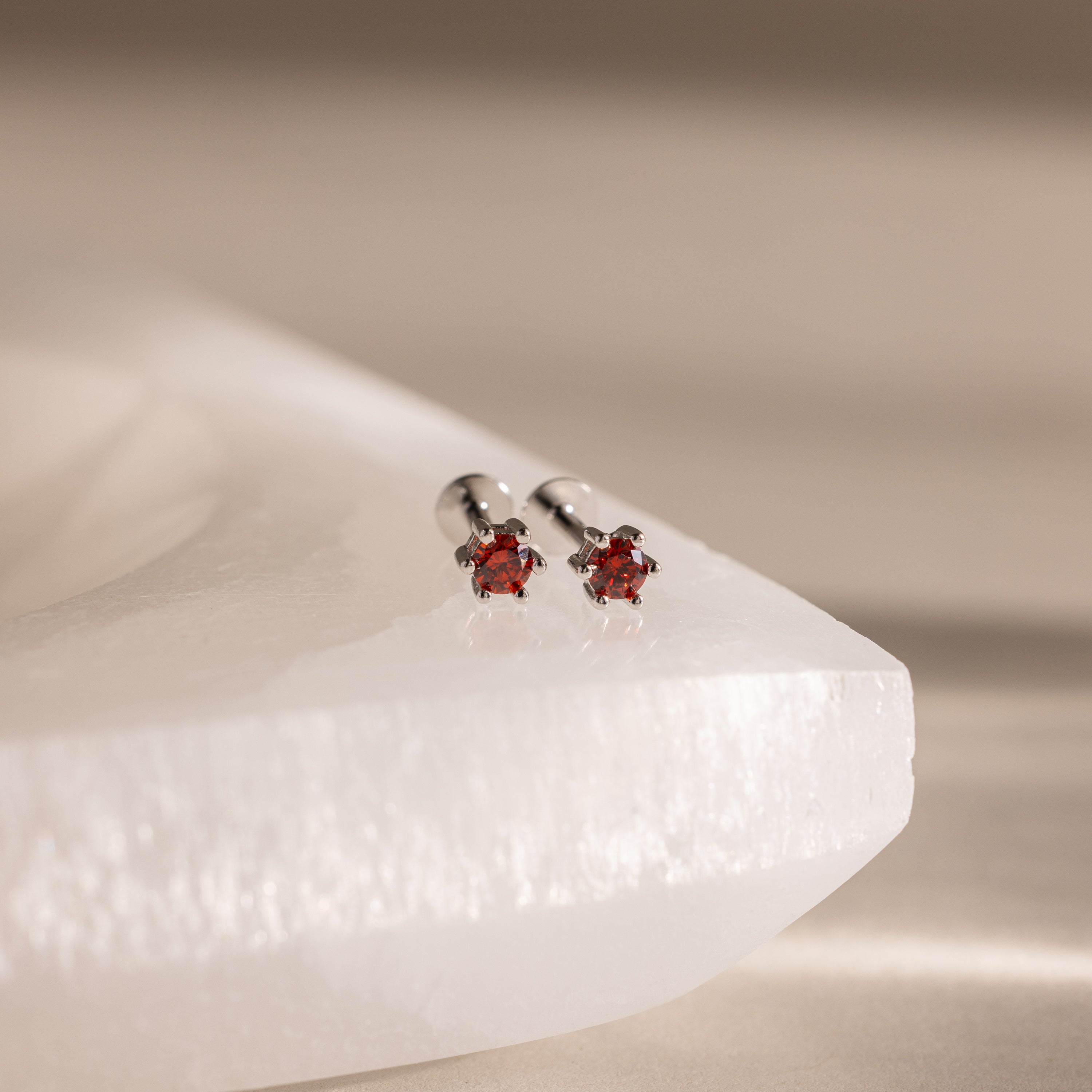 The Garnet Flatback Studs are two silver stud earrings with round red gemstones, perfect as birthstone studs or dainty earrings, shown on a white textured surface.
