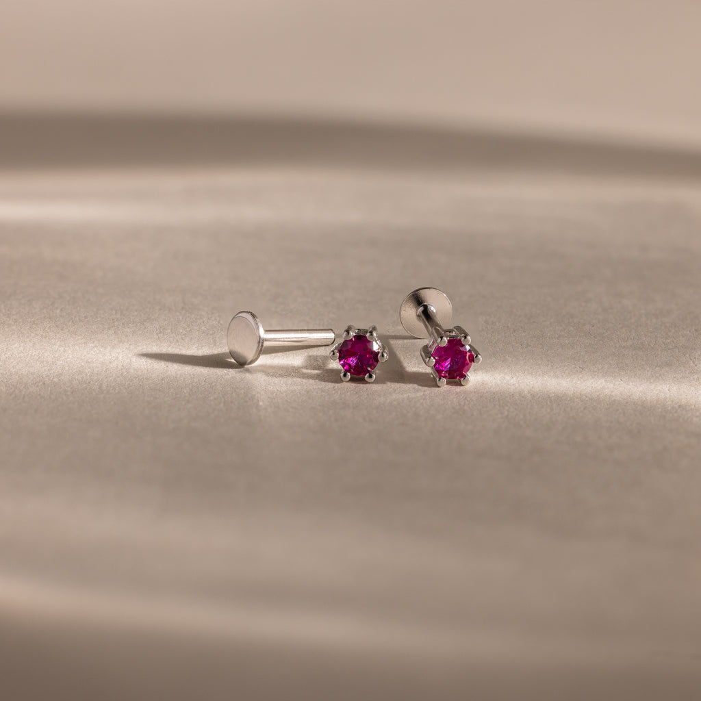 The Birthstone Flatback Studs are silver earrings featuring small round red gemstones, designed as elegant birthstone studs and displayed on a beige surface.