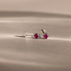The Birthstone Flatback Studs are silver earrings featuring small round red gemstones, designed as elegant birthstone studs and displayed on a beige surface.