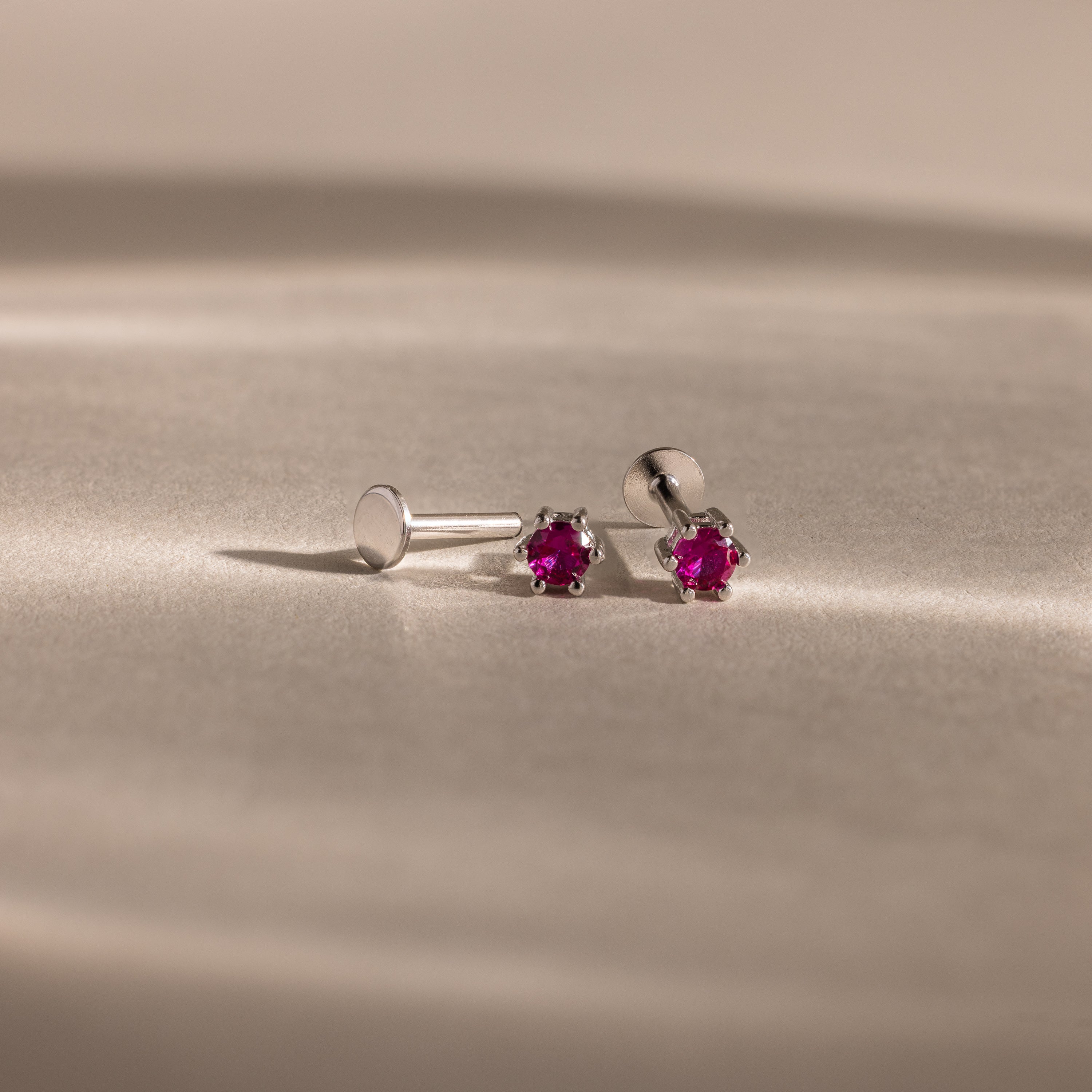 The Birthstone Flatback Studs are silver earrings featuring small round red gemstones, designed as elegant birthstone studs and displayed on a beige surface.