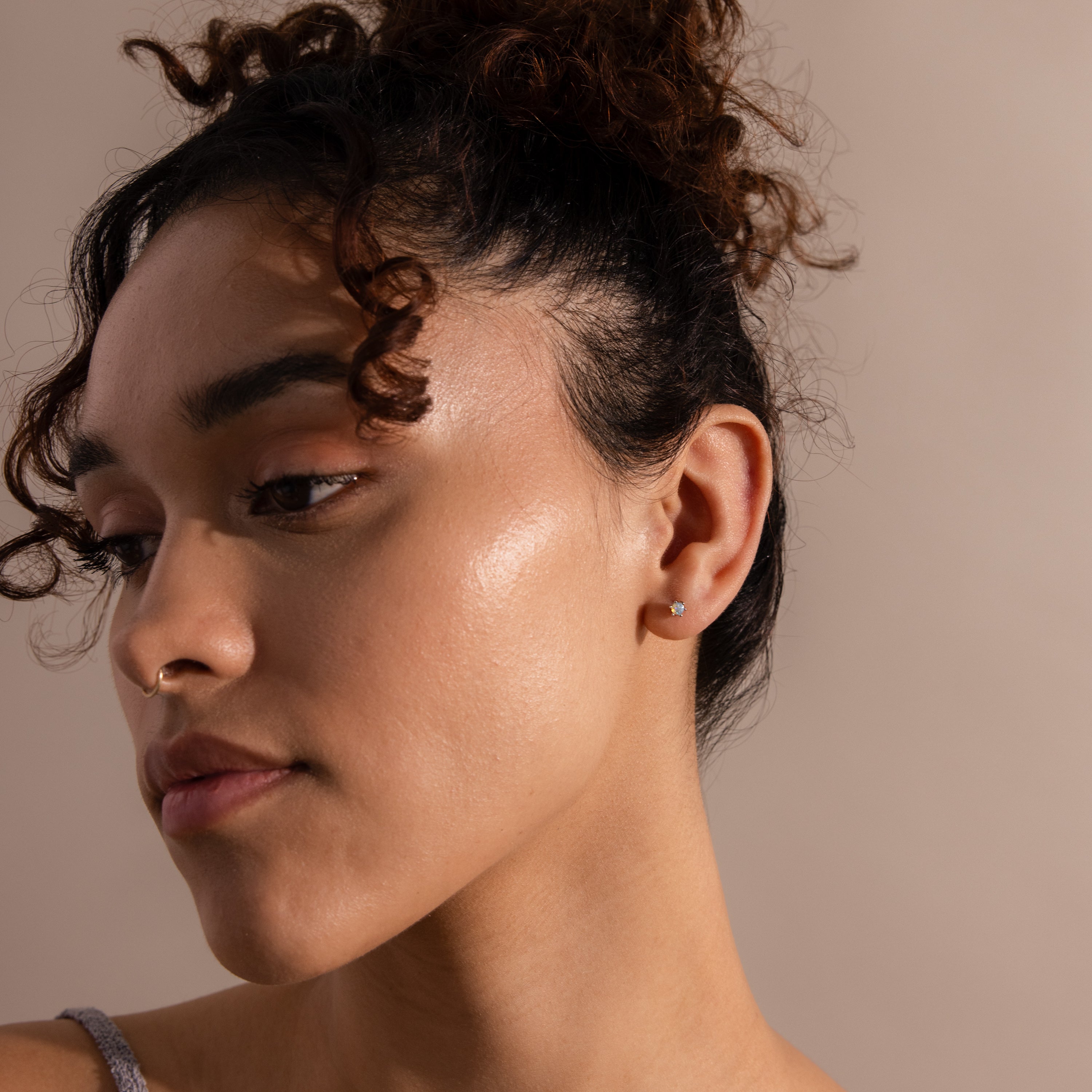 A woman with curly hair in a bun, wearing a nose ring and subtle Opal Flatback Studs, looks down against a neutral background.