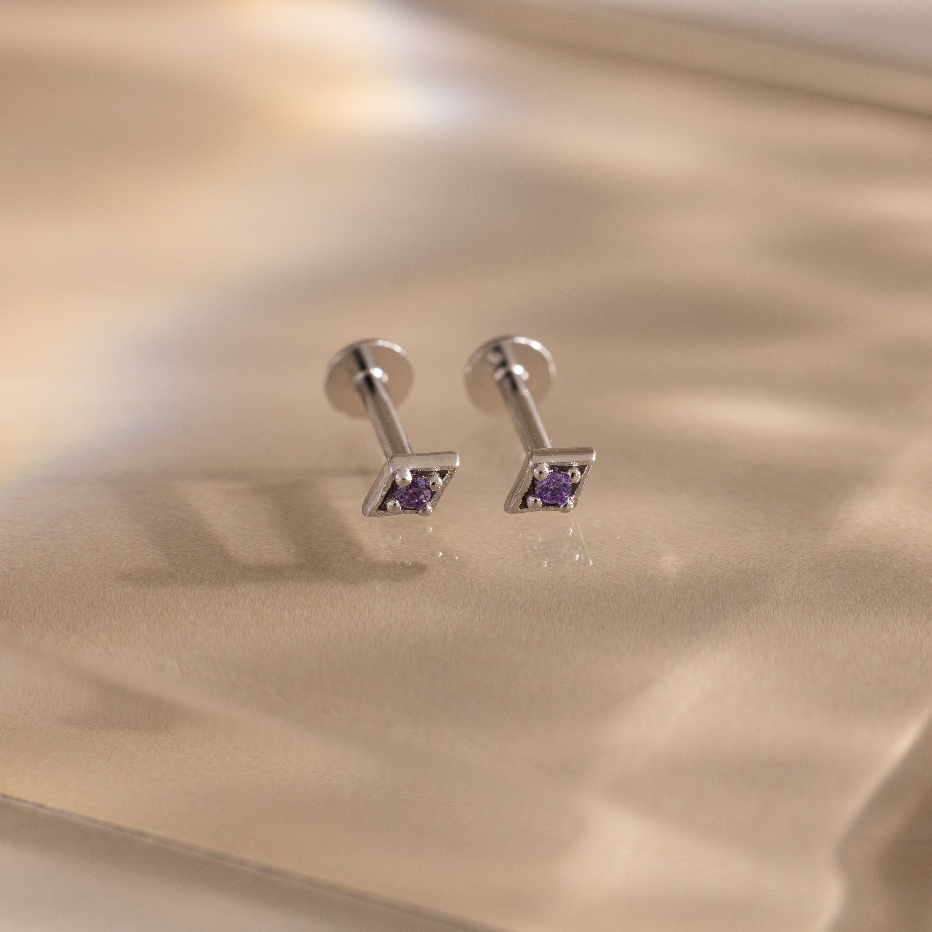 Pair of silver geometric flat back amethyst stud earrings on a beige fabric background