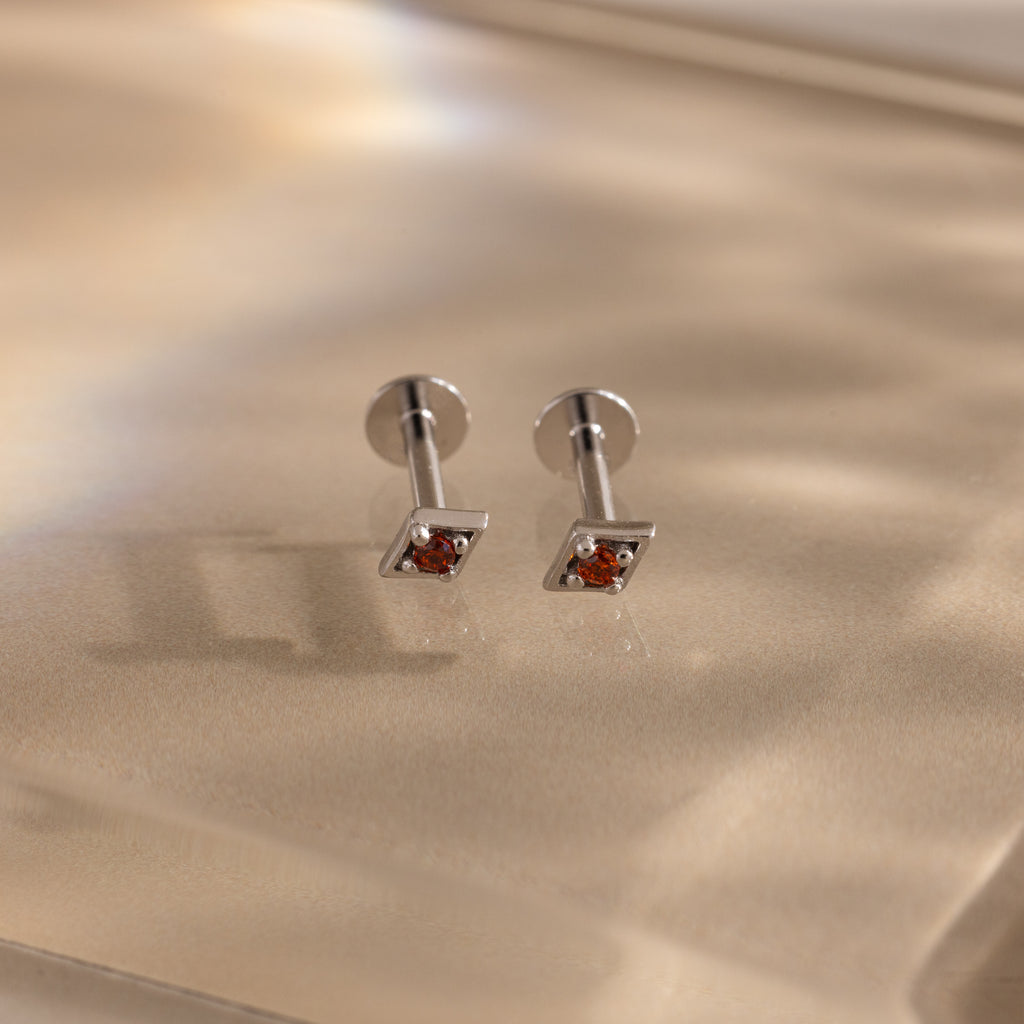 The Geometric Garnet Flat Back Studs feature small red gemstones set in silver, shown on a soft beige surface.