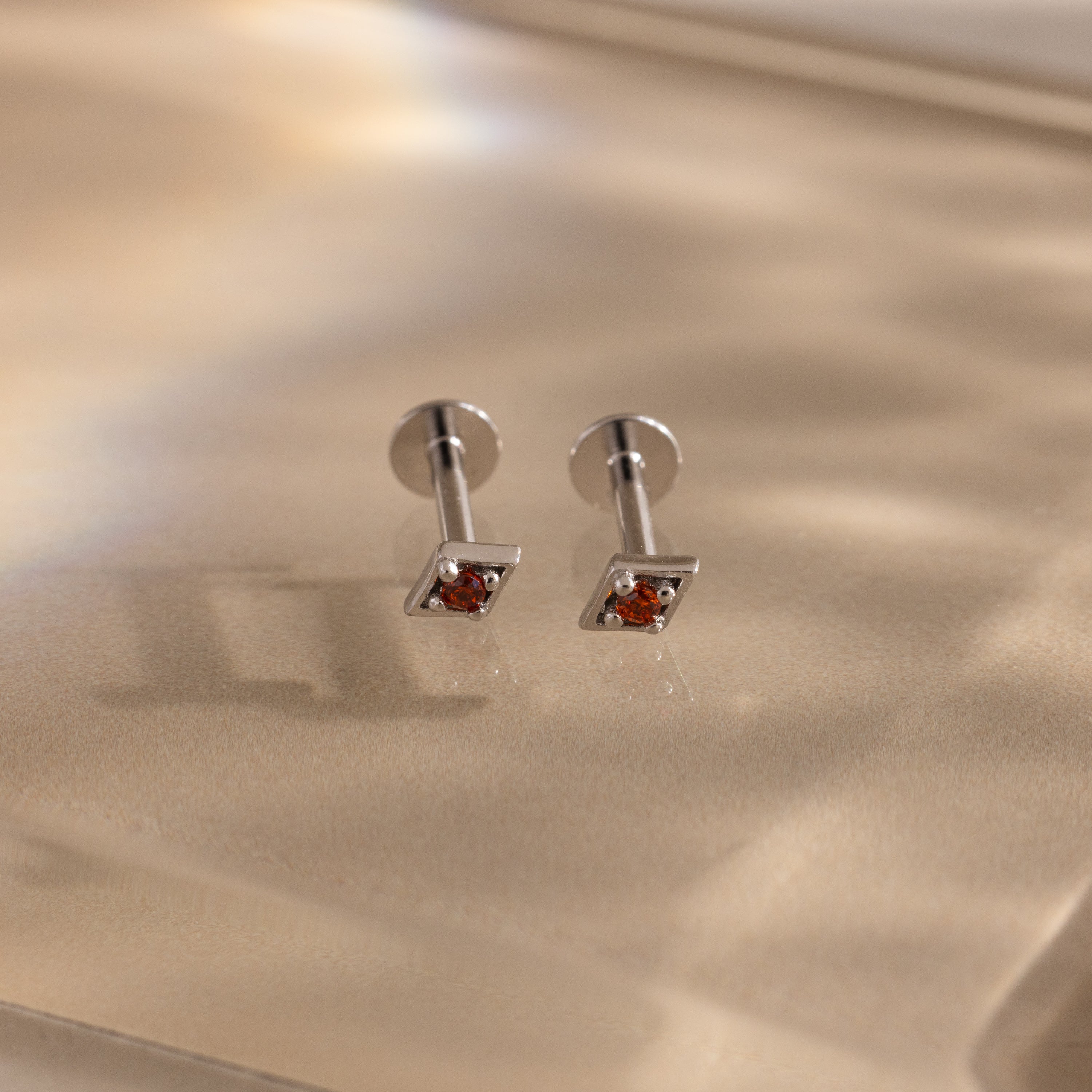 The Geometric Garnet Flat Back Studs feature small red gemstones set in silver, shown on a soft beige surface.