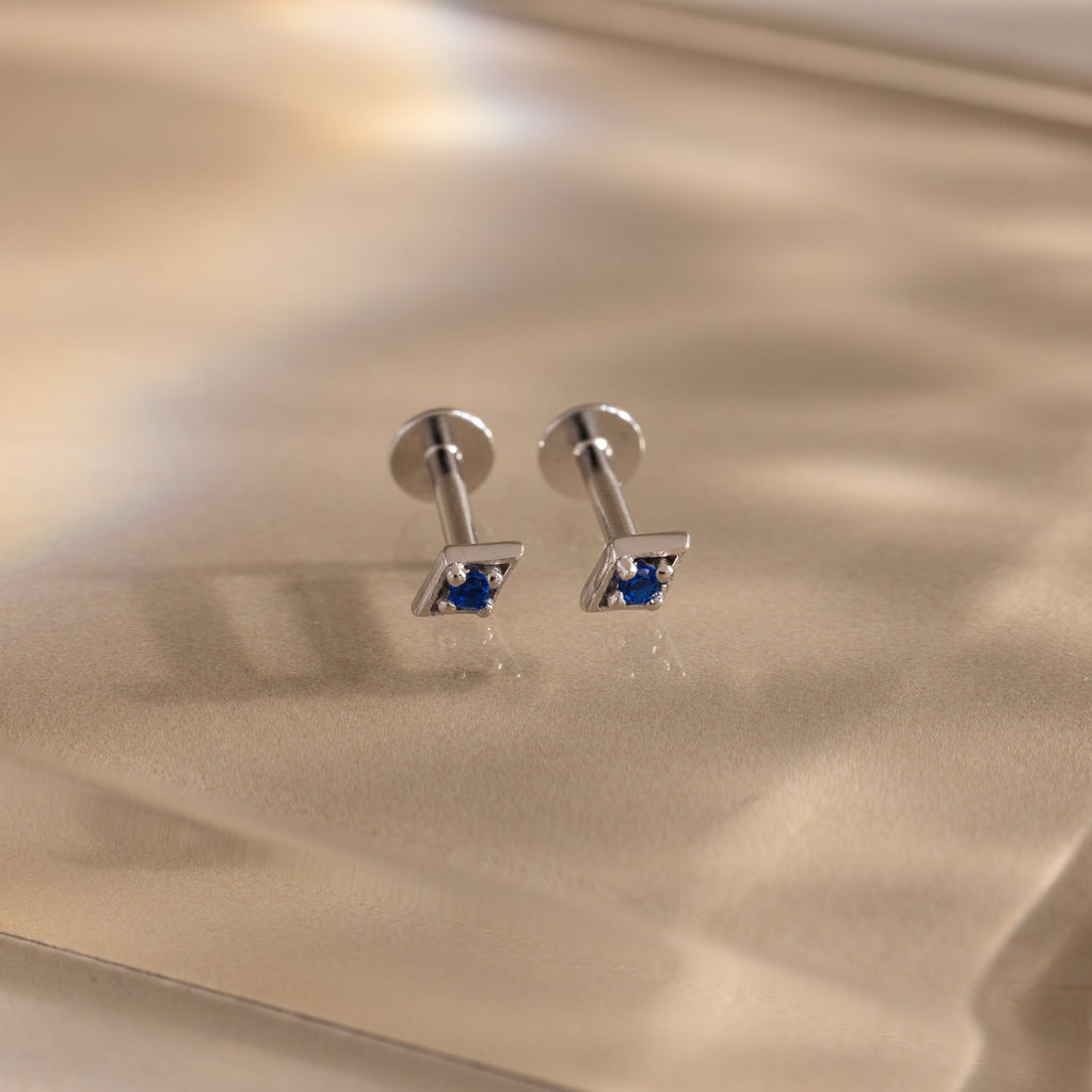 Pair of silver geometric flat back sapphire stud earrings on a reflective gold surface