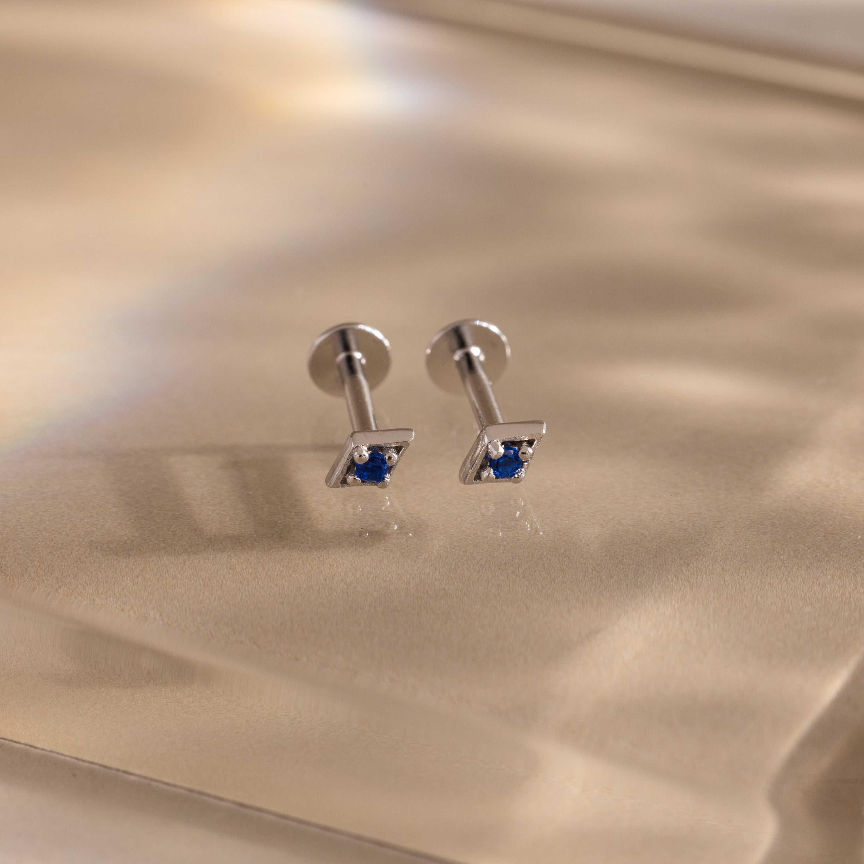 Pair of silver geometric flat back sapphire stud earrings on a reflective gold surface