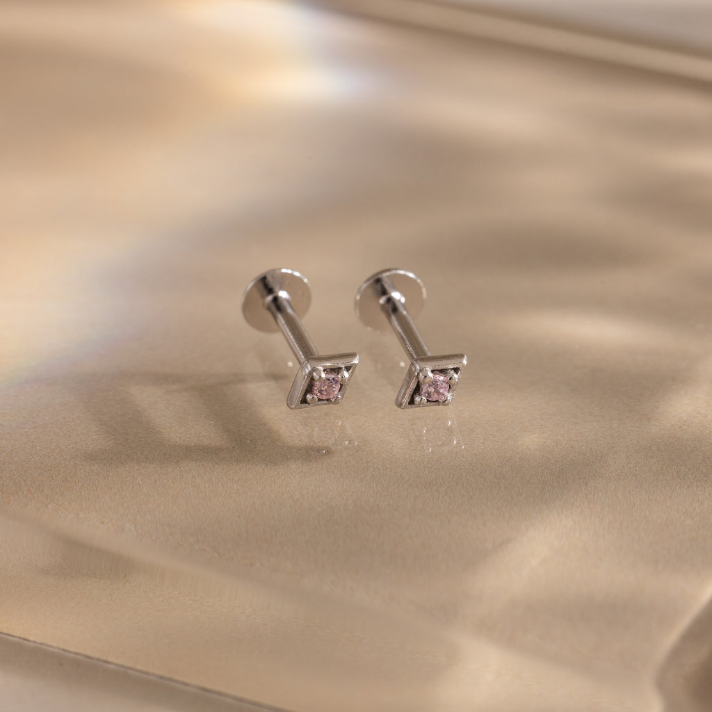 The Geometric Pink Tourmaline Flat Back Studs, featuring silver settings and geometric pink tourmaline gemstones, rest on a soft beige surface for an elegant birthstone earring look.