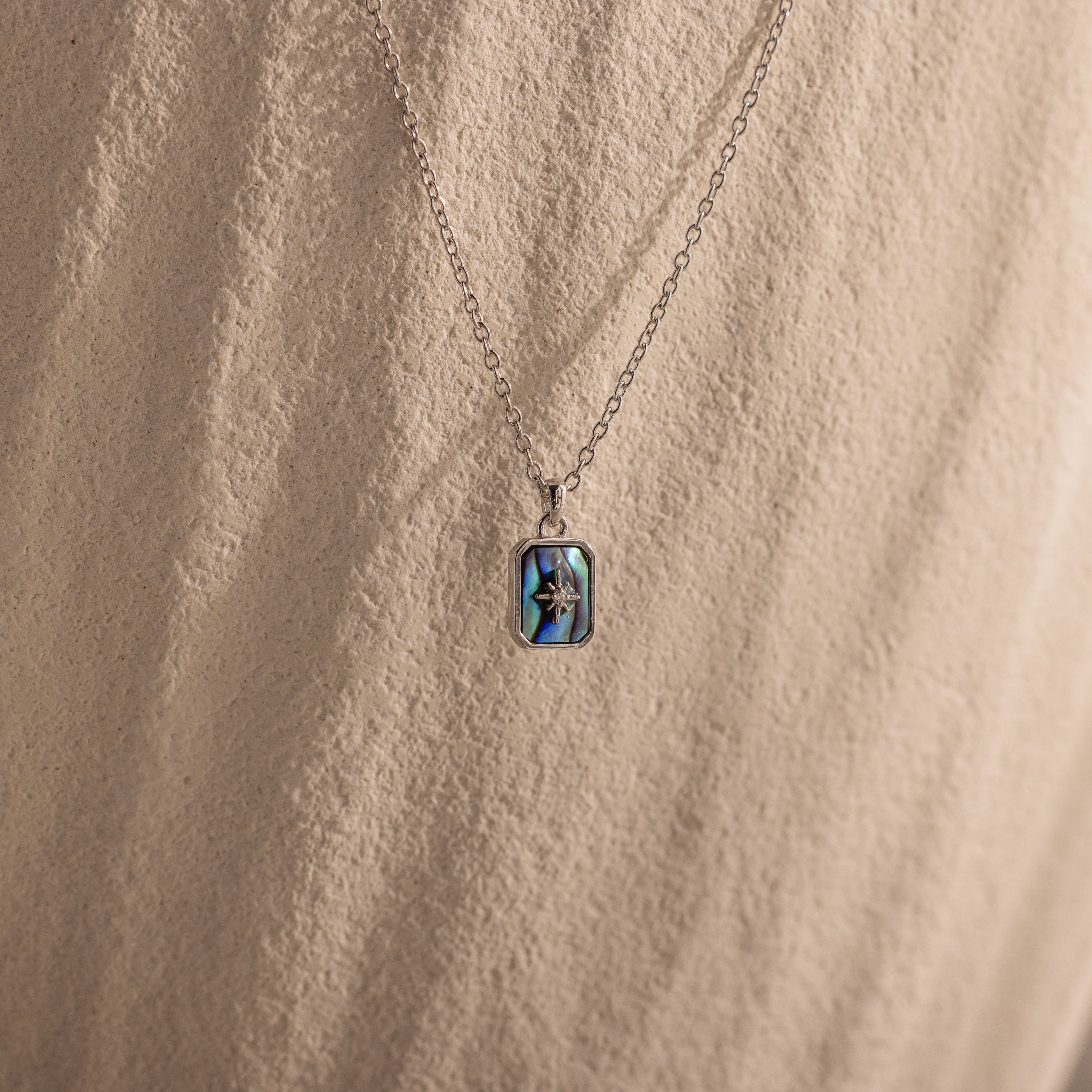 The North Star Gemstone Necklace in Sterling Silver features an abalone rectangular pendant, resembling a crystal, displayed on a textured surface.
