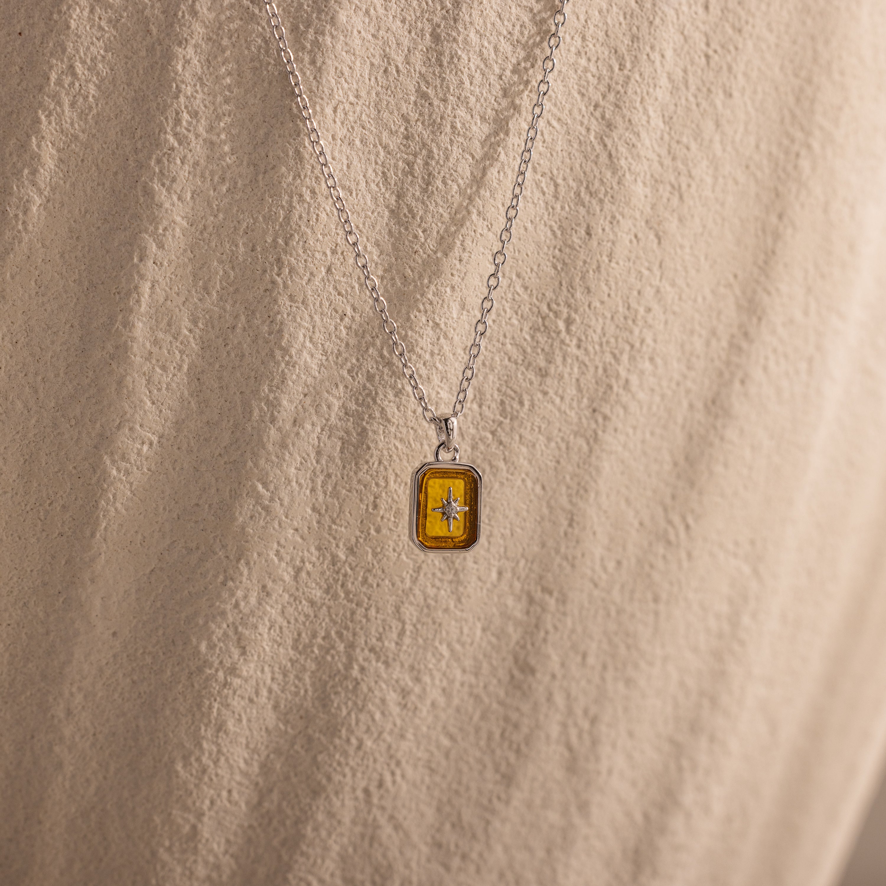 The North Star Topaz Necklace, featuring a silver chain and a rectangular topaz pendant, is displayed on a textured beige background.