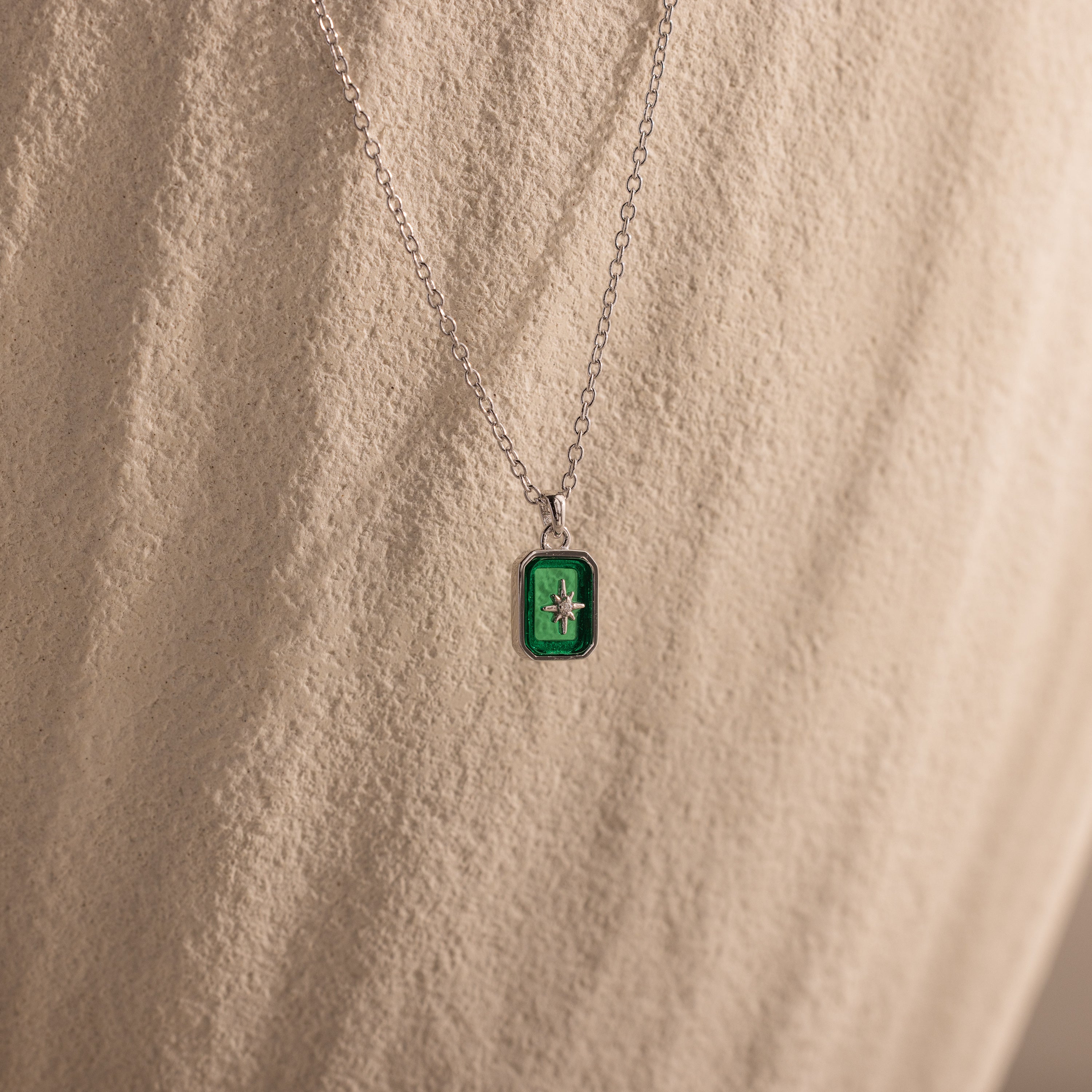 The North Star Emerald Necklace features a green rectangular crystal pendant with a delicate star design, showcased on a textured surface.