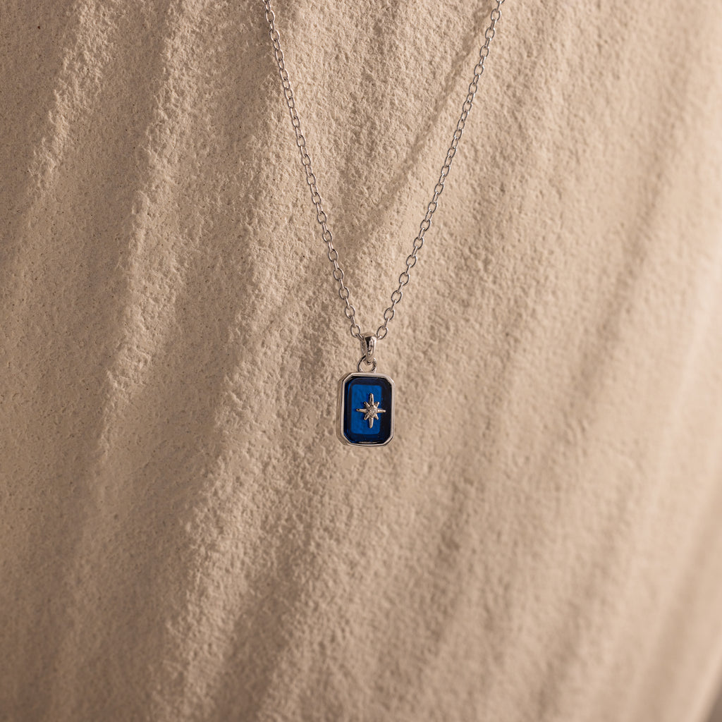 The North Star Sapphire Necklace features a blue rectangular crystal pendant with a diamond-set North Star, displayed on a textured beige surface.