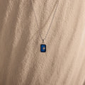 The North Star Sapphire Necklace features a blue rectangular crystal pendant with a diamond-set North Star, displayed on a textured beige surface.
